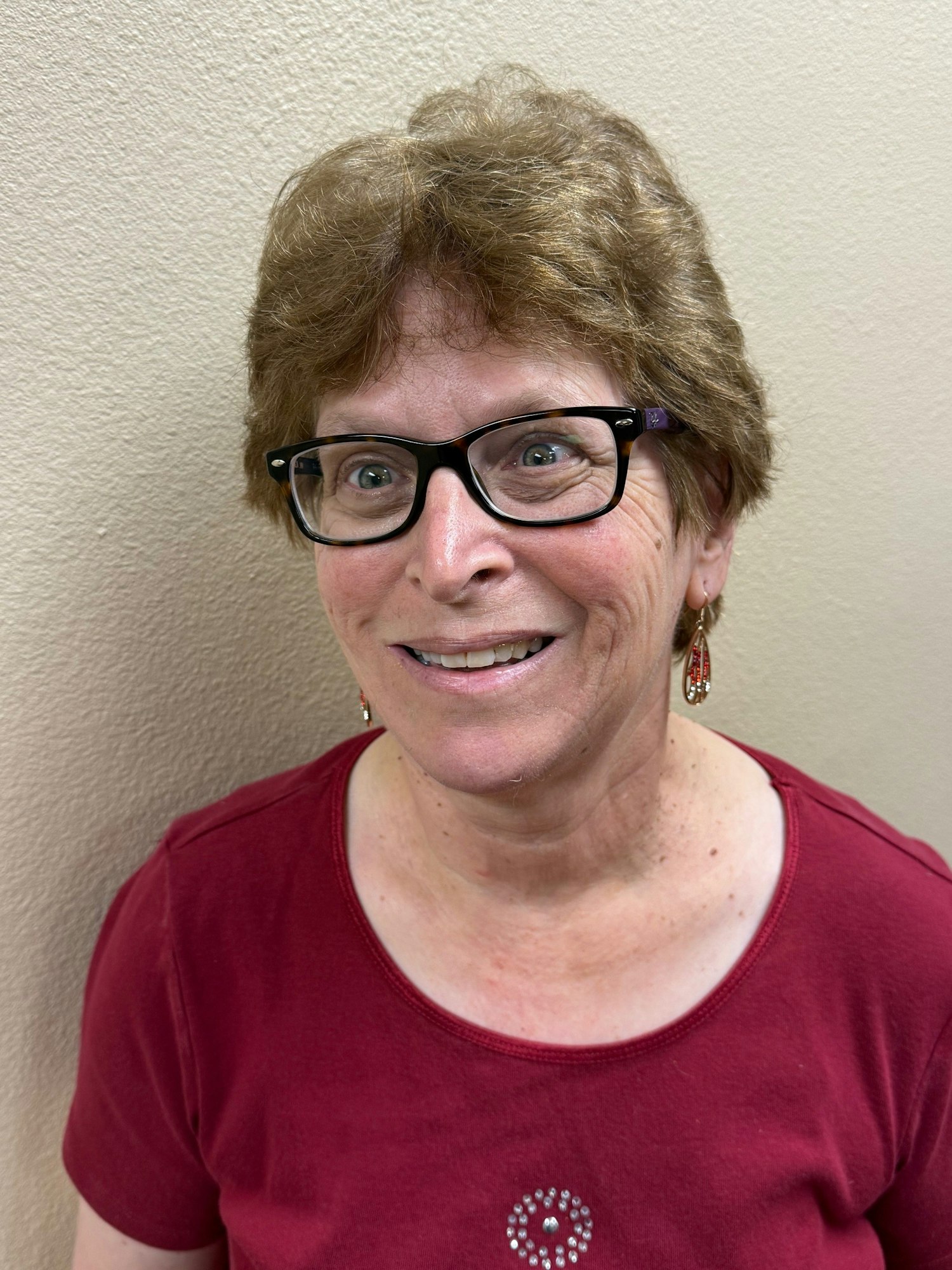 A person wearing glasses and a red shirt with a pattern, smiling in front of a light-colored wall.