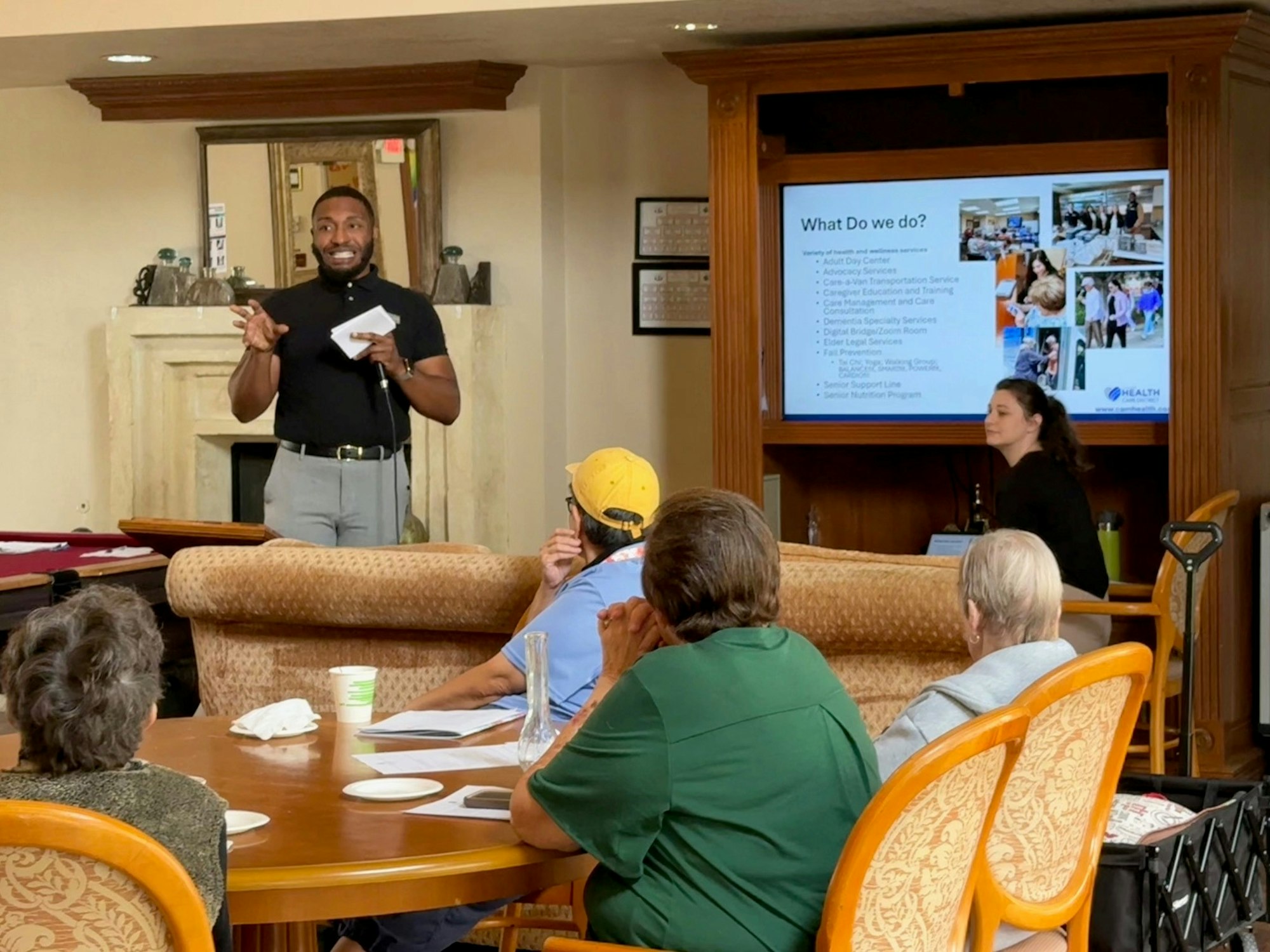 A speaker engages with an audience in a community setting, discussing services for seniors, while a presentation displays information.