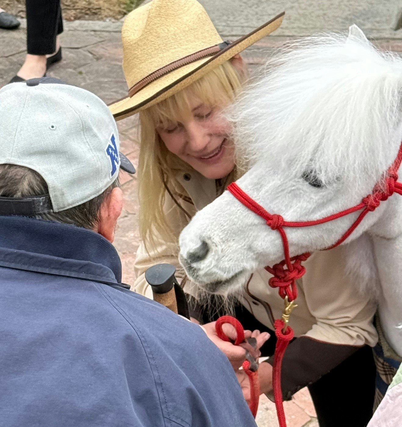A woman in a hat with a white pony, speaking to a man in a cap outdoors.