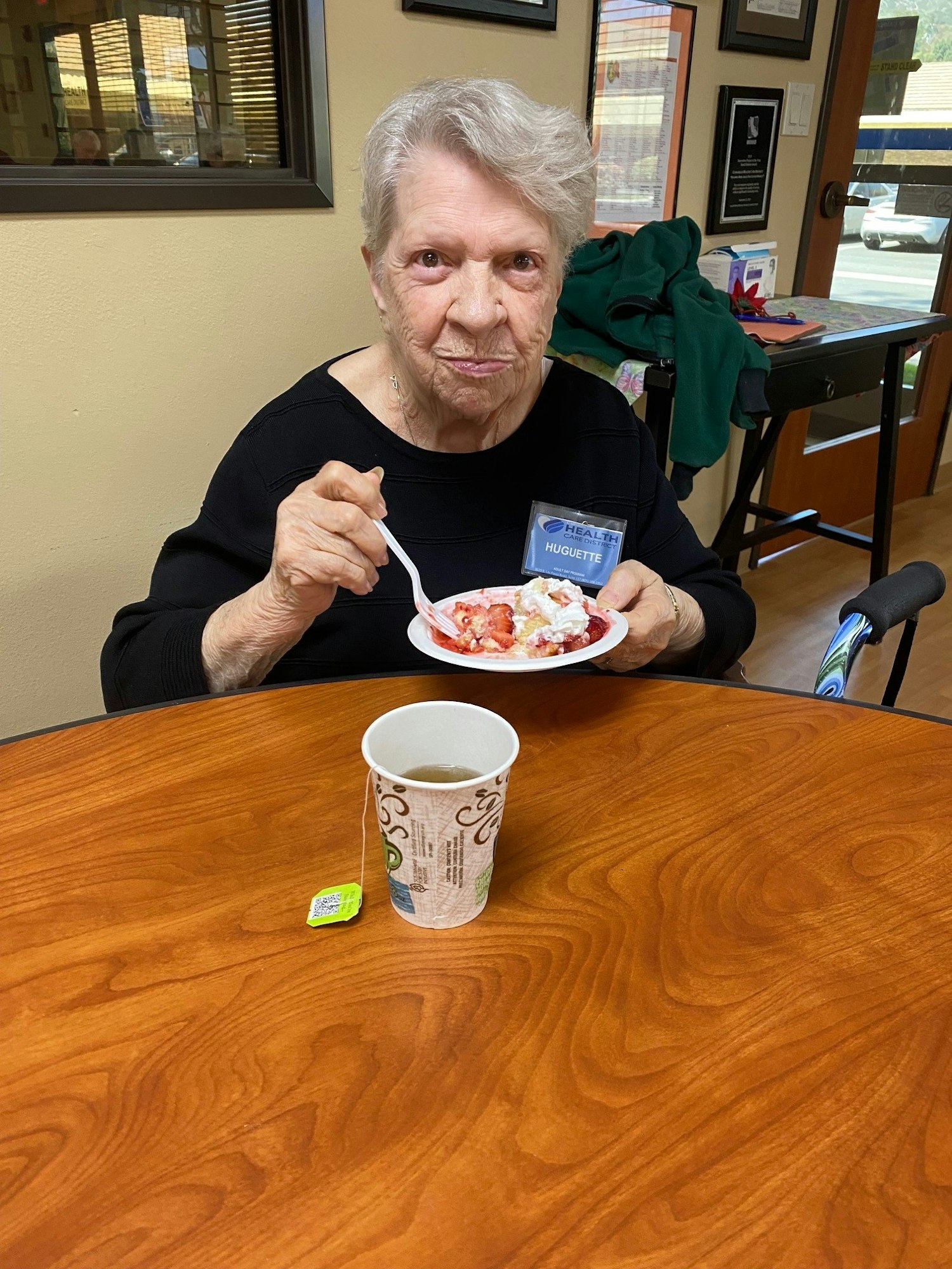 Elderly person eating dessert at a table with a cup of tea and a green tea bag nearby.