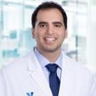 A person wearing a white lab coat and blue tie, smiling, with a blurred background.