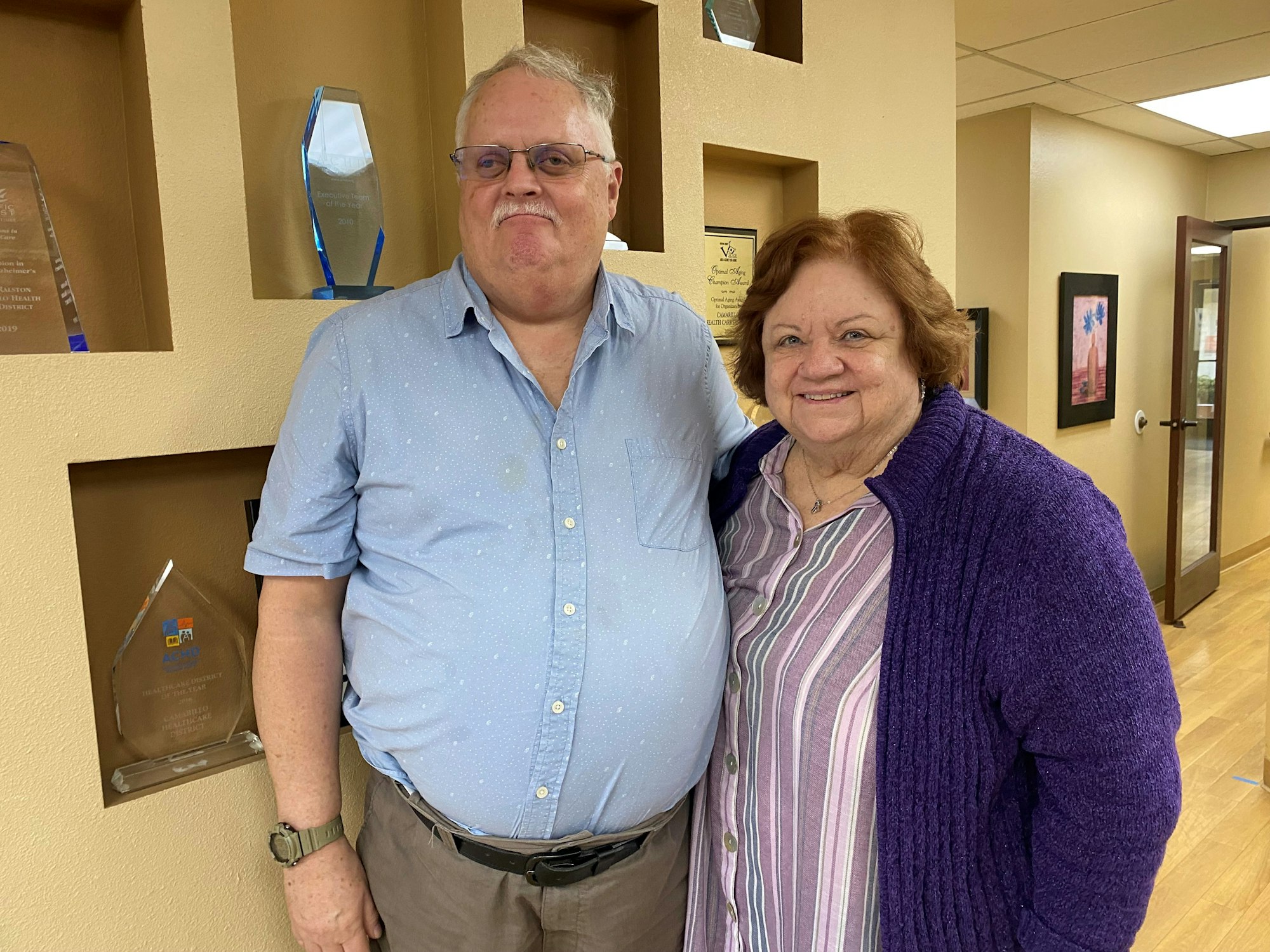 Two people standing inside, with awards displayed on the wall behind them.