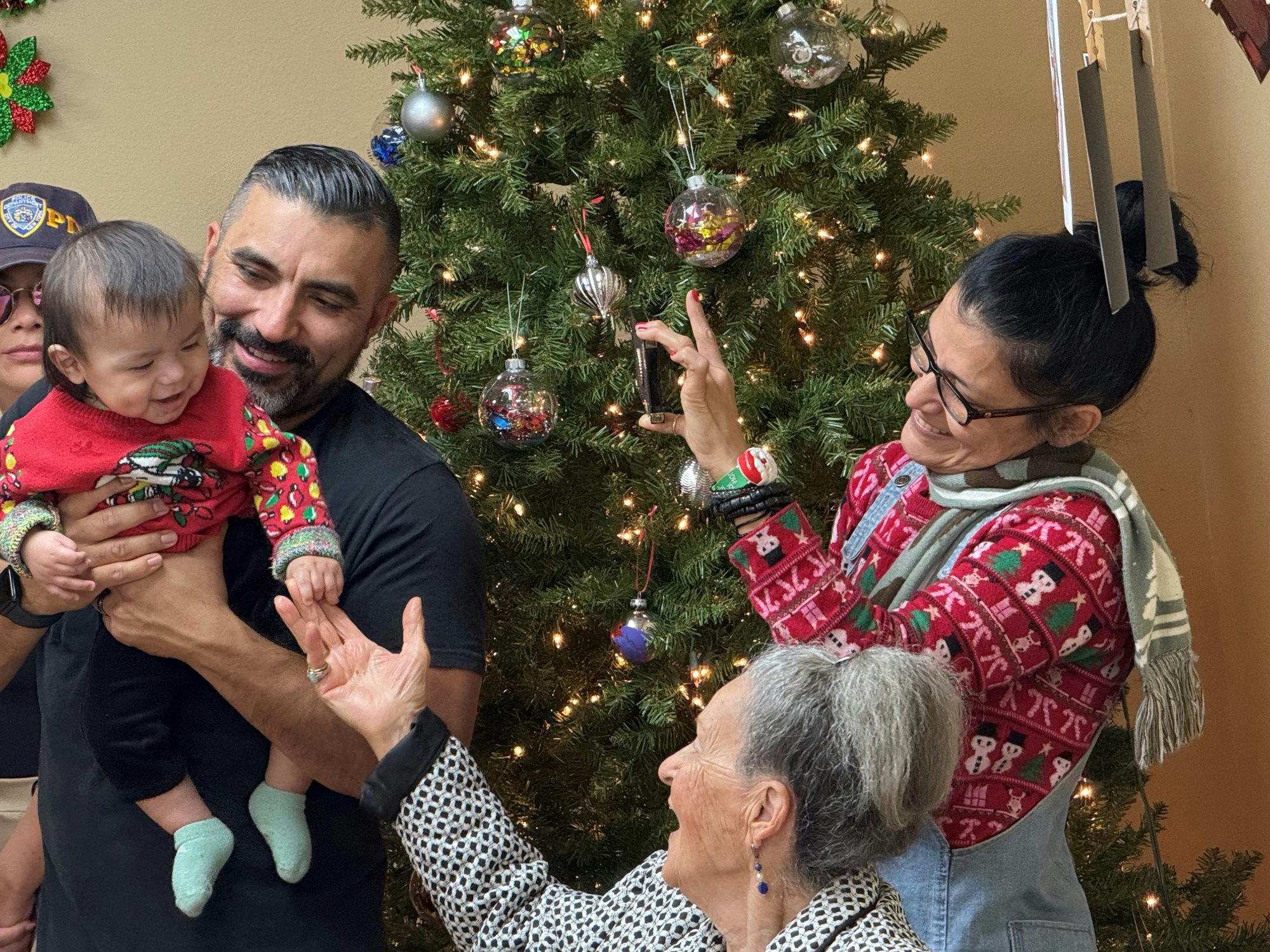 A family enjoys decorating a Christmas tree, with a happy baby in a festive sweater being held.