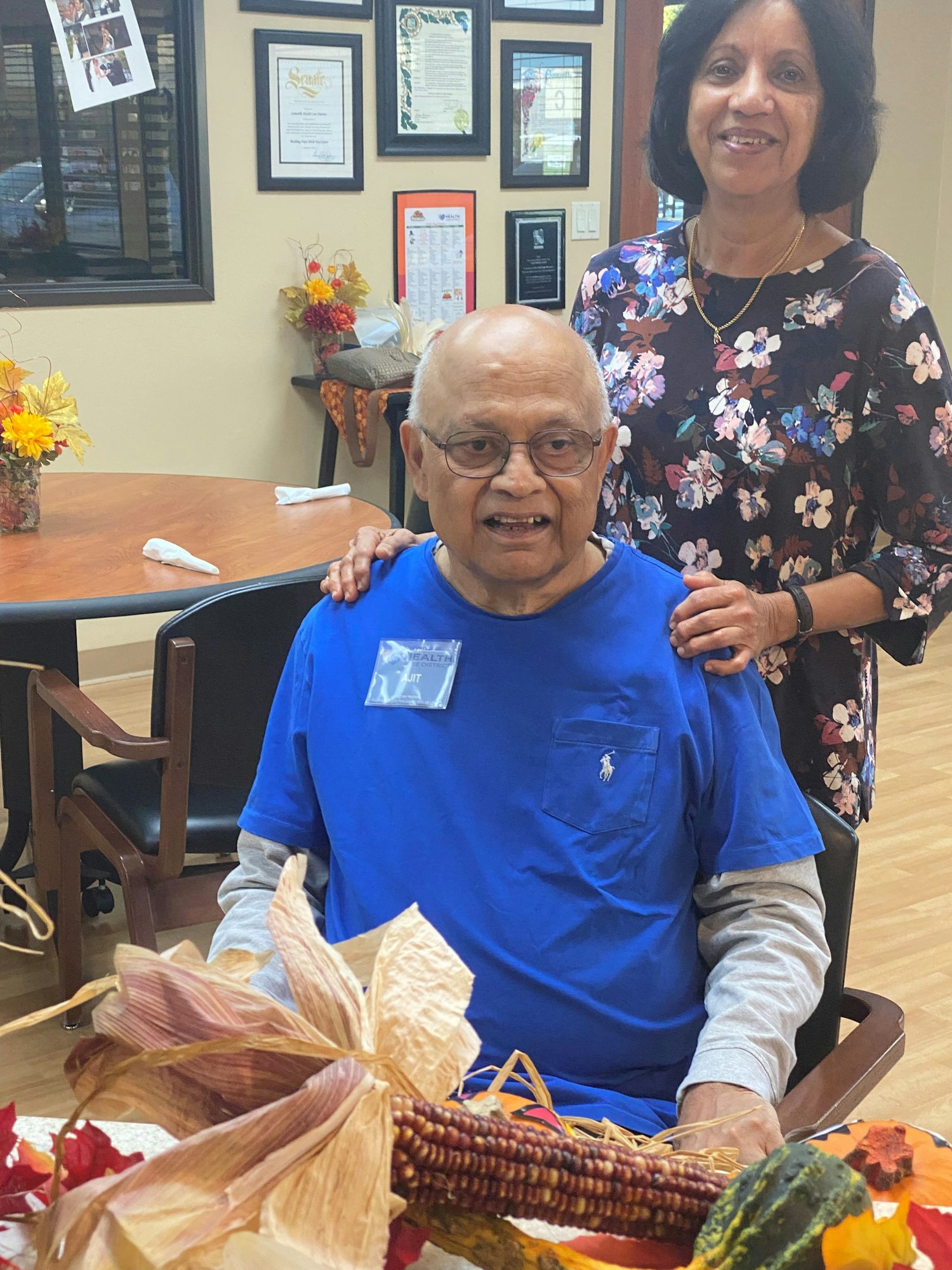 A smiling elderly man in a blue shirt is seated at a table, with a woman behind him, in a warm, welcoming environment.