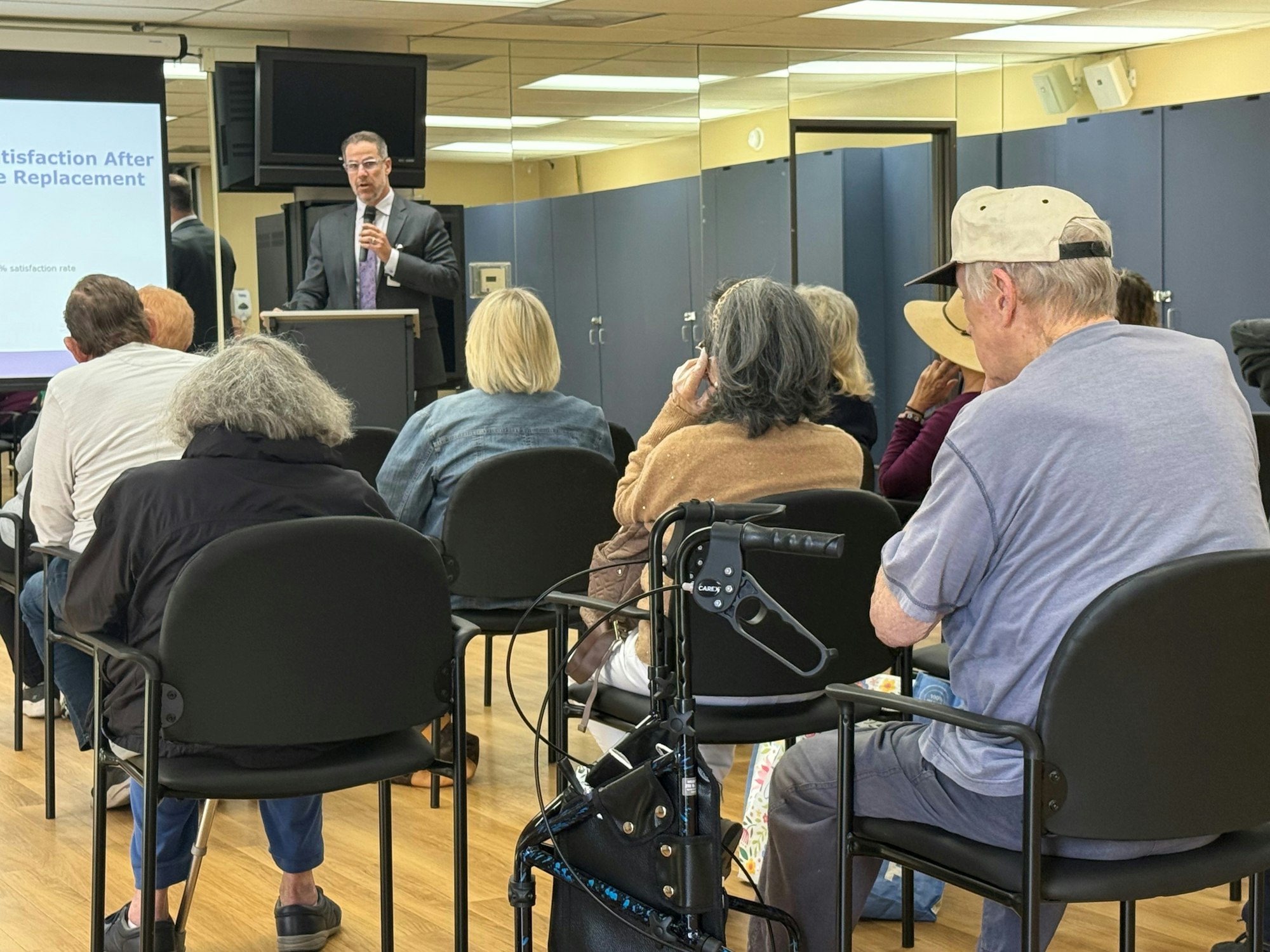 A speaker is addressing an audience seated indoors, with a presentation on knee replacement visible on a screen.