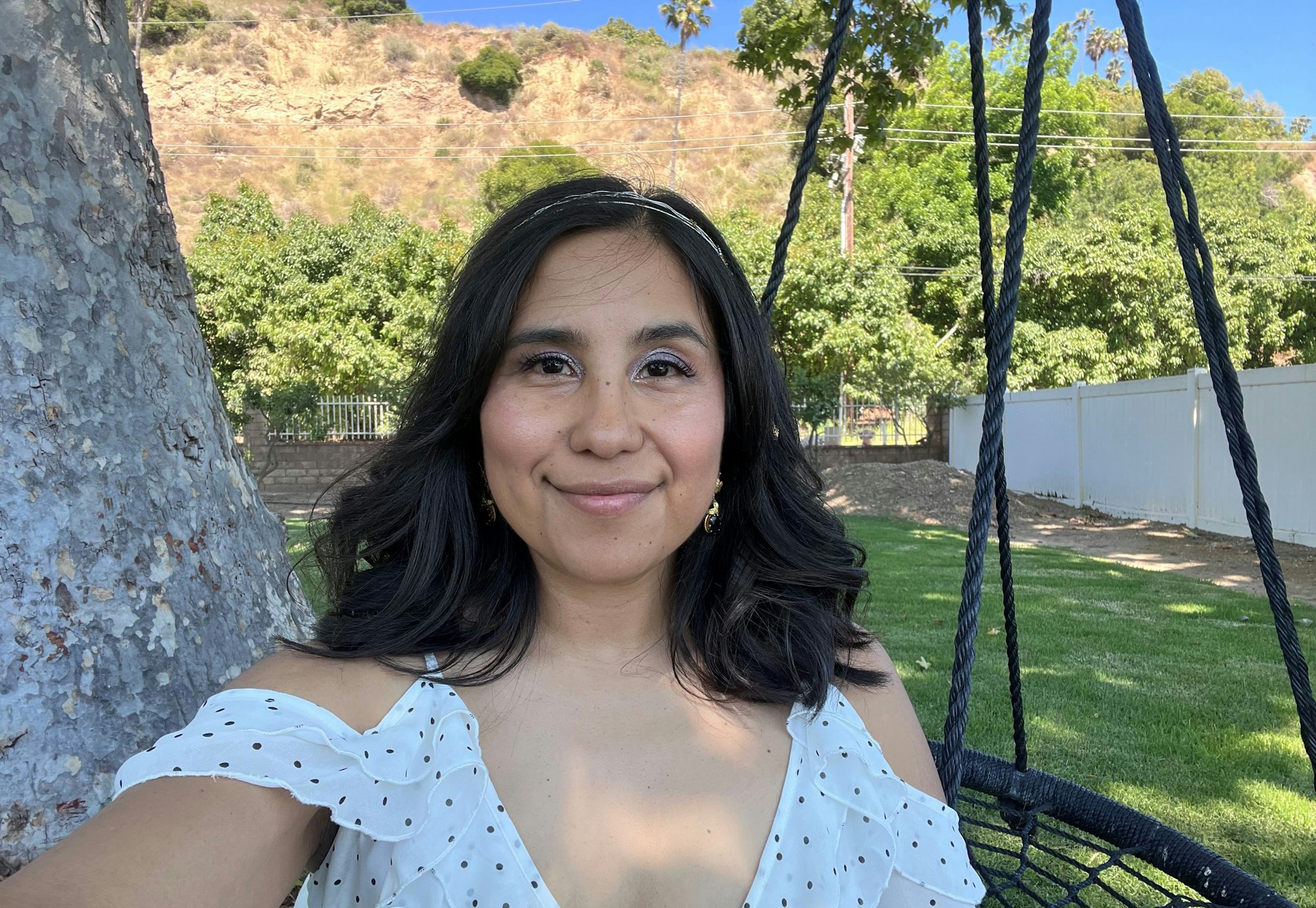 A woman in a white dress with polka dots is smiling outdoors near a tree and a swing.