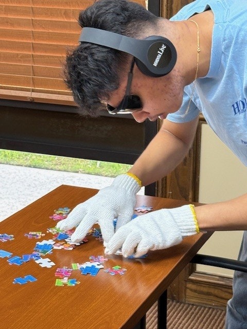 A person wearing headphones, sunglasses, and gloves attempts to assemble a puzzle on a table.