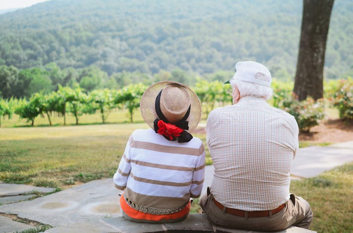An elderly couple sitting on a stone path, enjoying a scenic view of vineyards and hills.