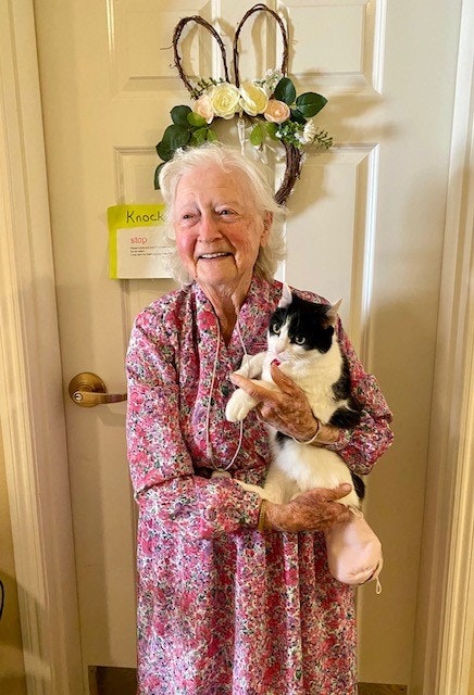Elderly woman in floral dress holding a black-and-white cat, standing by a decorated door.