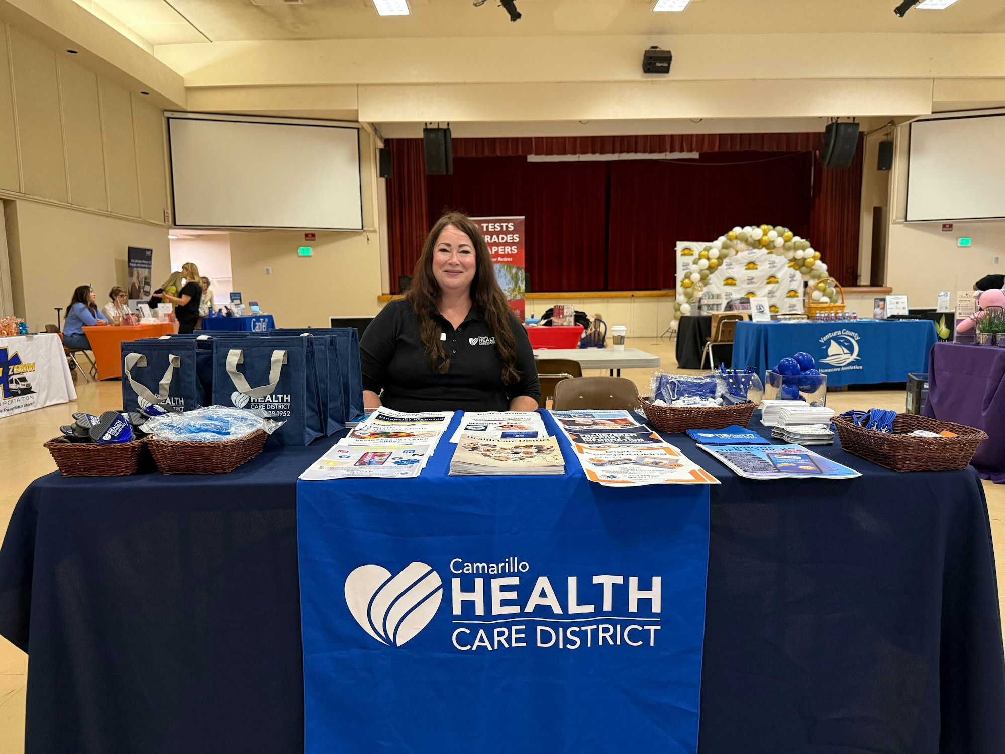 A woman at a Camarillo Health Care District table with brochures, tote bags, and giveaway items in a community event hall.