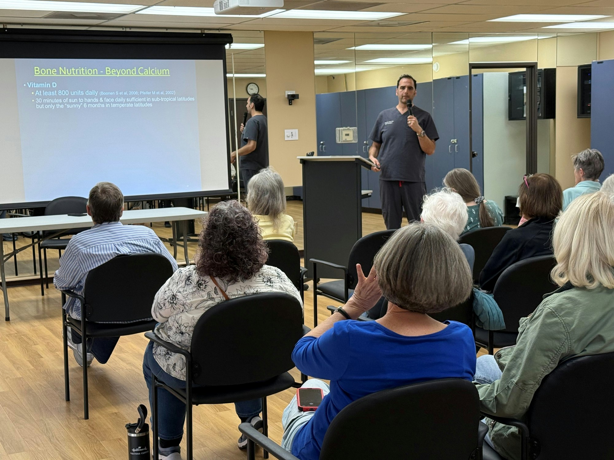 A speaker presents about bone nutrition, focusing on Vitamin D, to an audience seated at tables in a classroom-like setting.