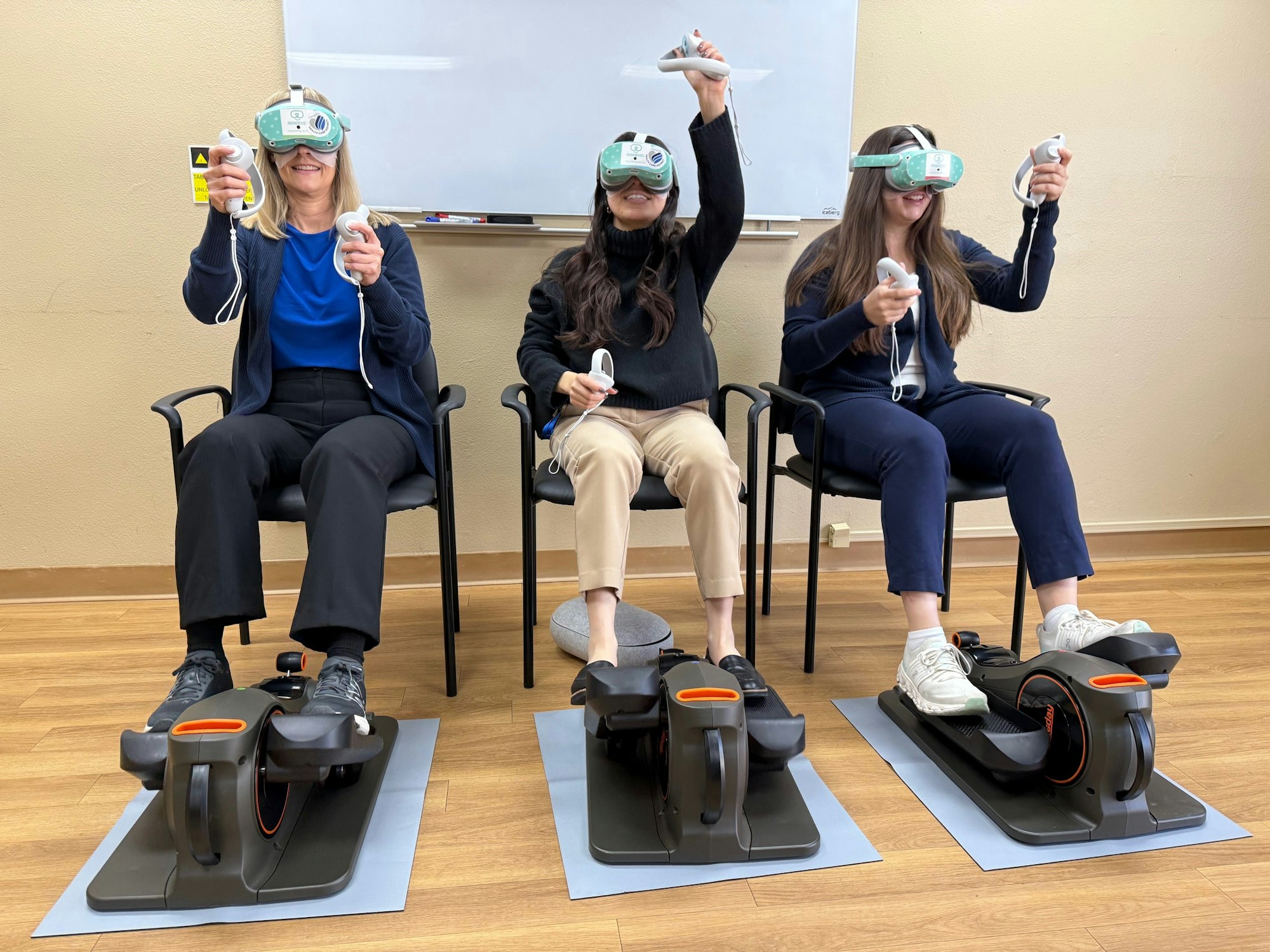 Three people using VR headsets and hand controllers, seated with feet in motion simulator devices.