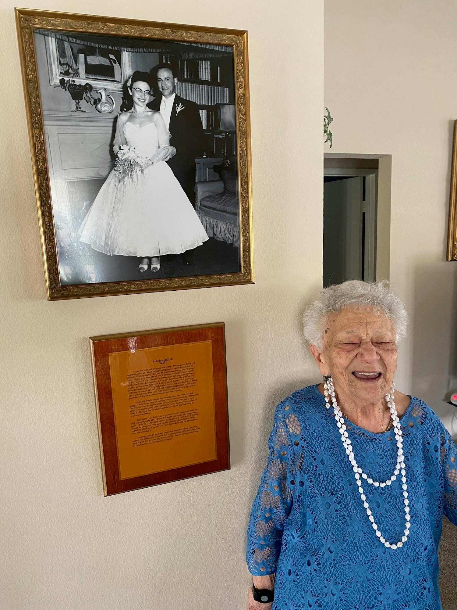 An elderly lady smiling, standing next to her framed wedding photo and a framed plaque on a wall.