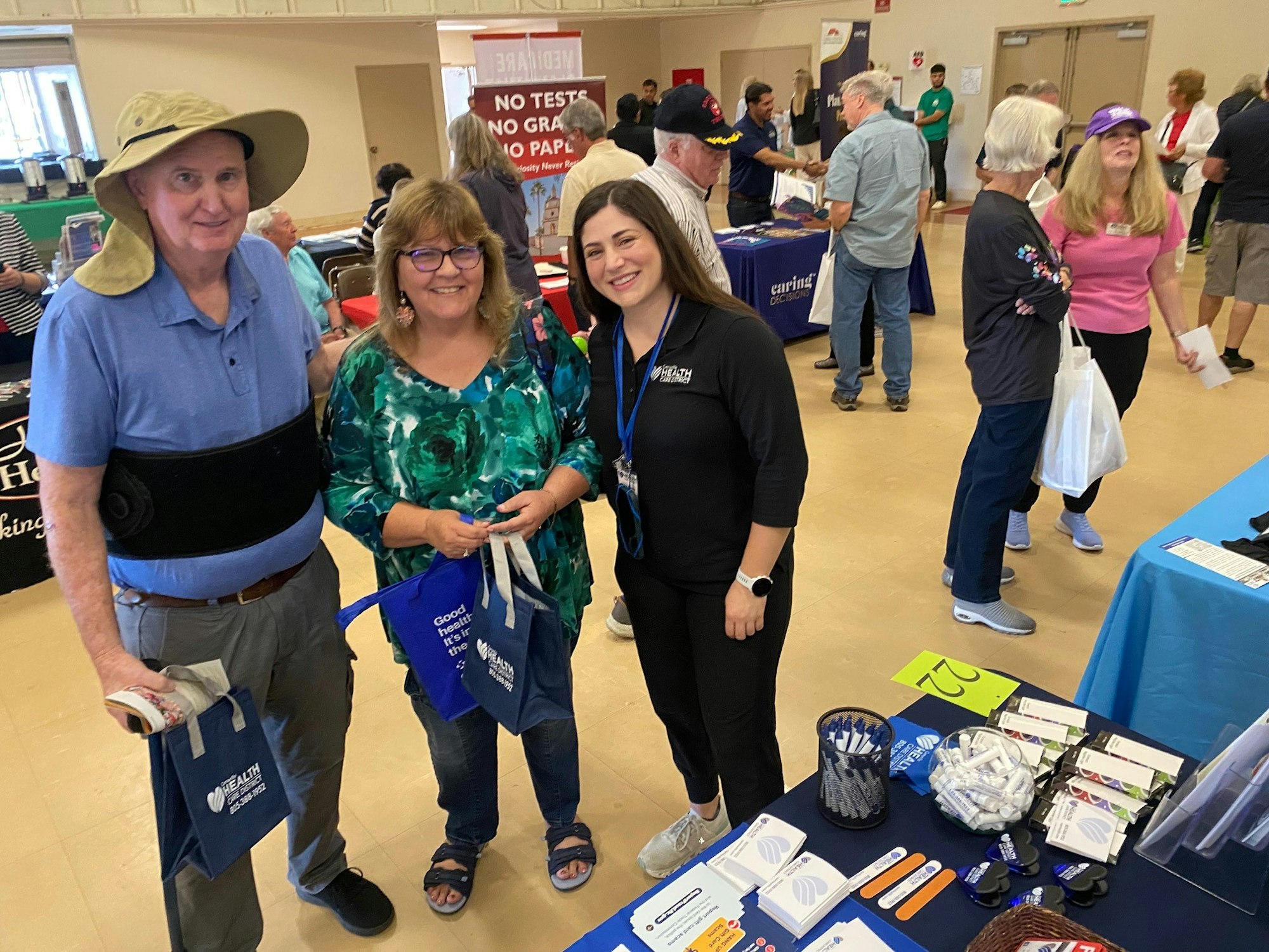 An indoor event with people at booths, promotional materials on a table, and attendees holding blue bags.