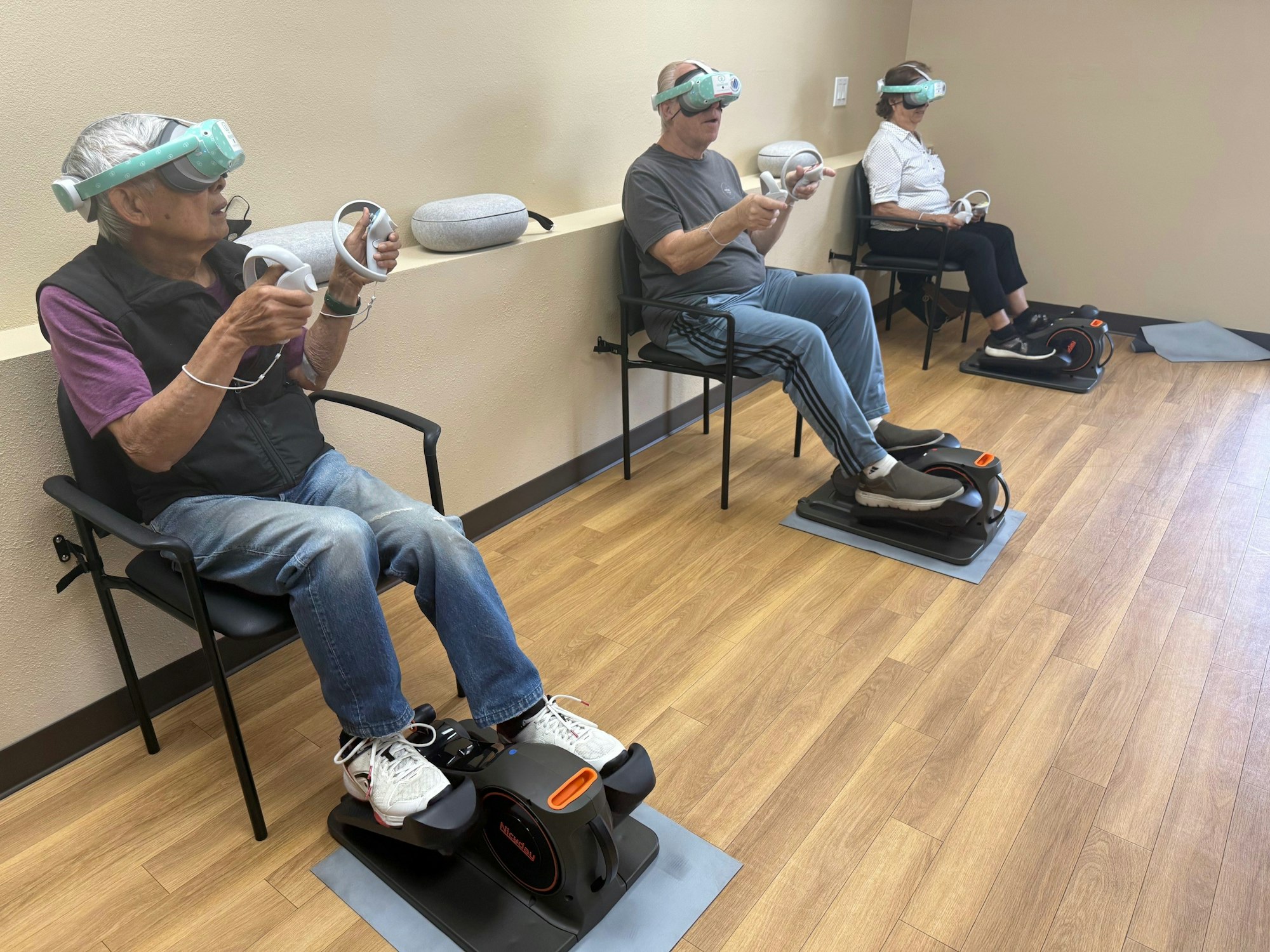 Three people wearing VR headsets and holding controllers, using seated exercise machines in a room.