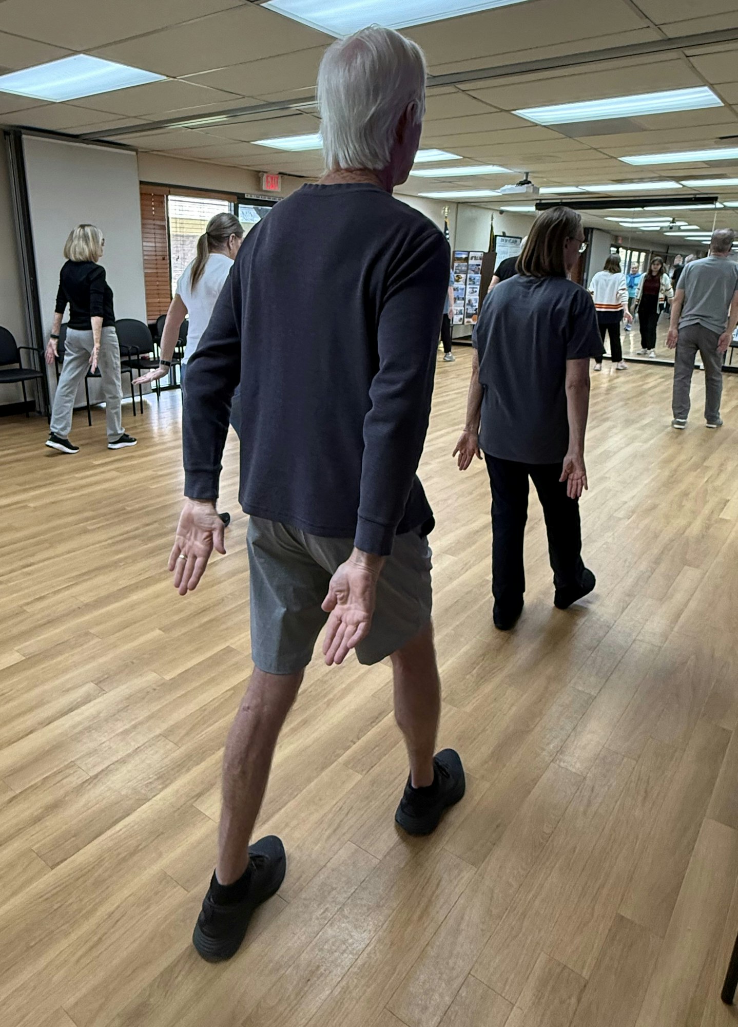 A group of people practicing movement or dance in a spacious room with wooden floors. Some have hands positioned behind them.