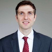 A man in a suit and red polka dot tie against a neutral background.