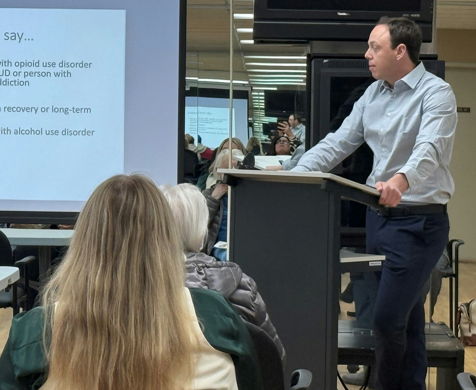 A man is giving a presentation on opioid use disorder to an audience in a room with a mirrored wall.
