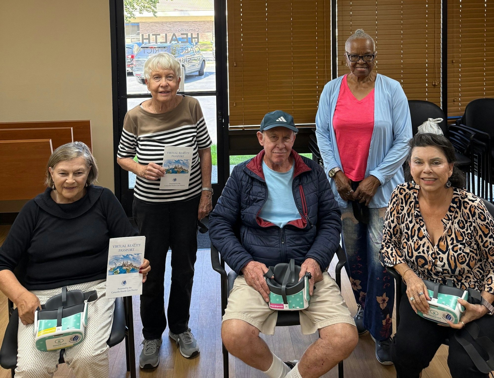 A group of five adults, some holding virtual reality headsets and a pamphlet, gather in a room with a glass door and chairs.