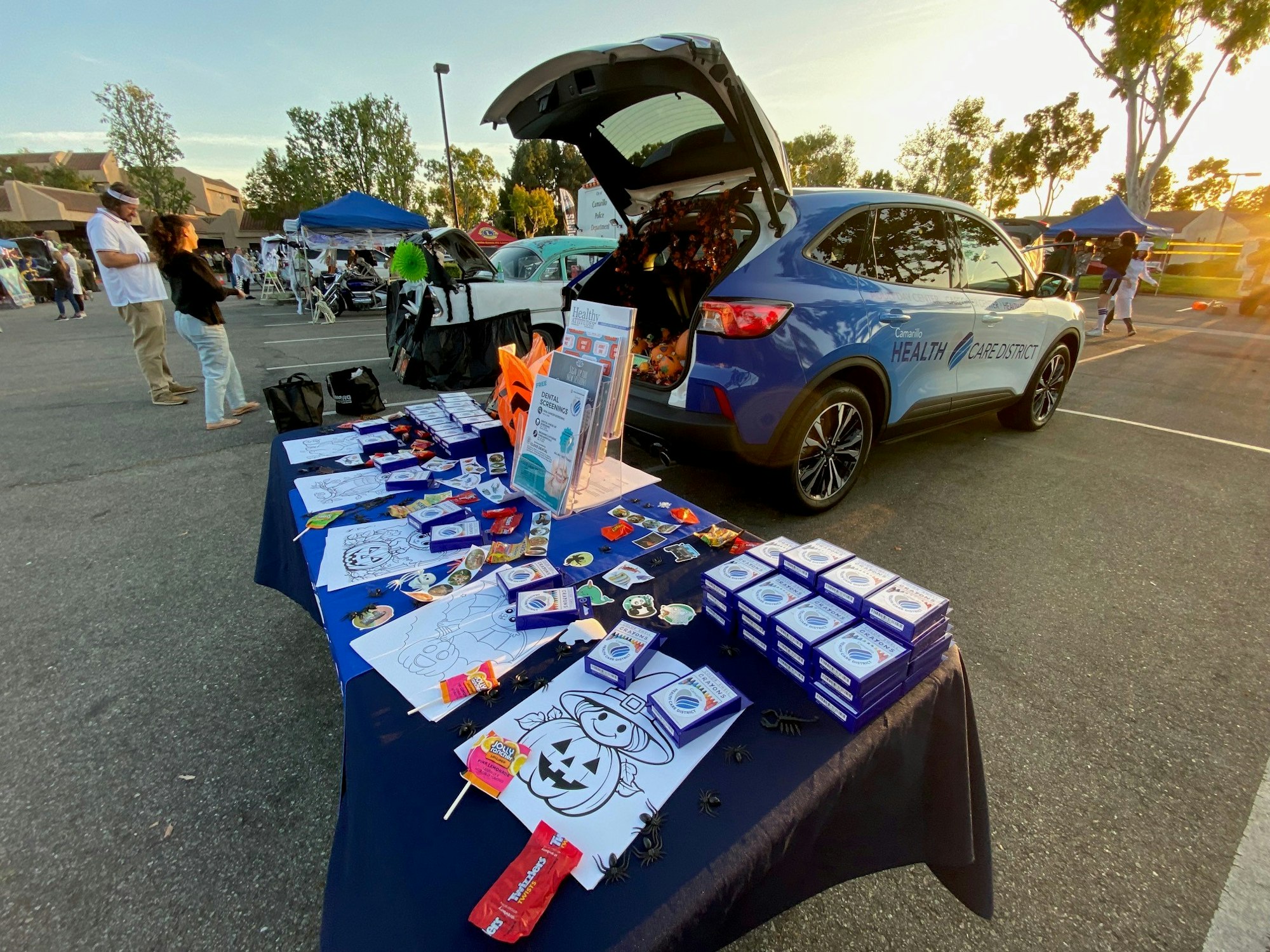Outdoor event with a car trunk open, a table with coloring pages, candy, and supplies, and people milling around nearby.