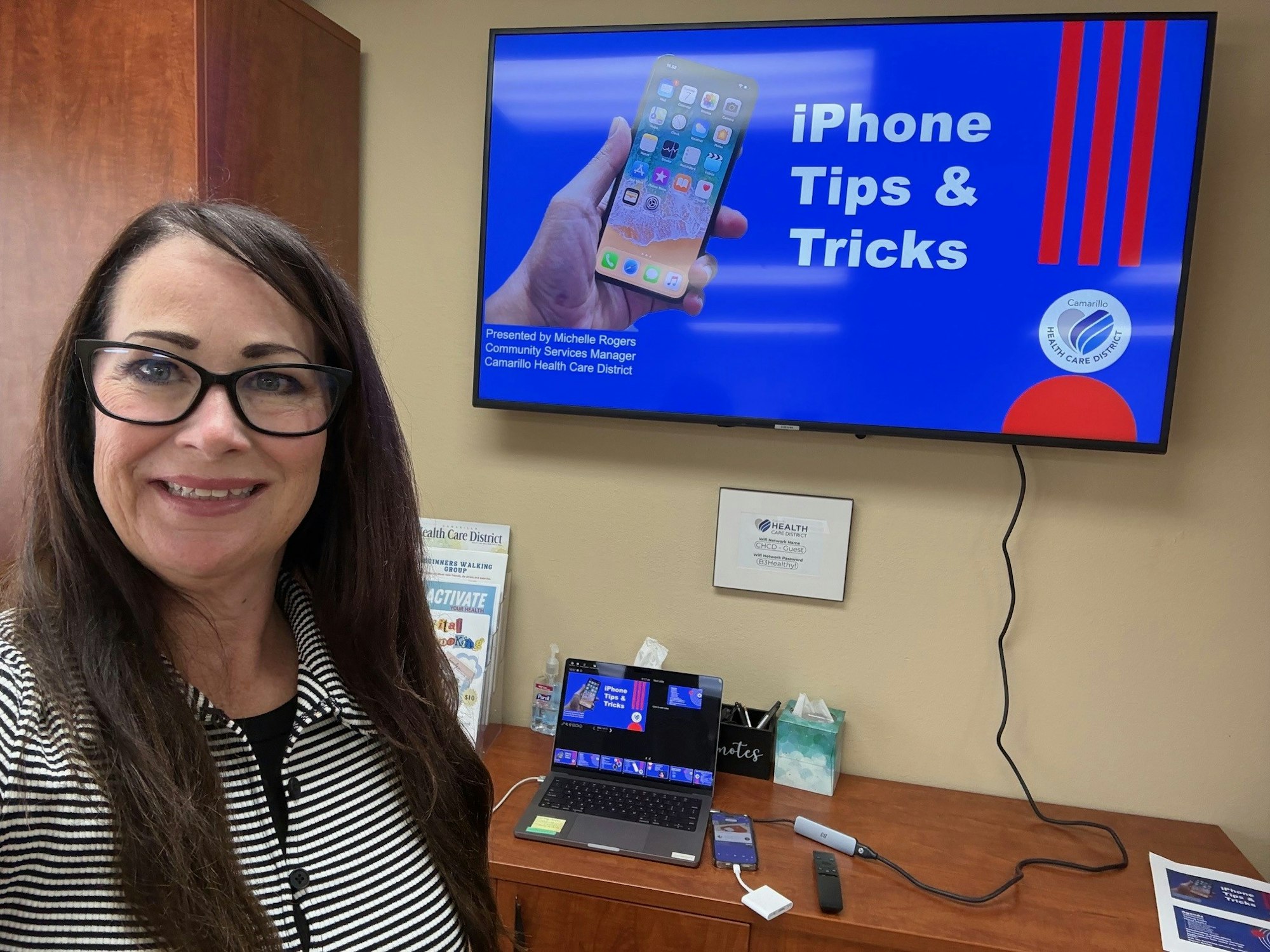 A woman presents an "iPhone Tips & Tricks" workshop, with a display screen and laptop set up in an office.
