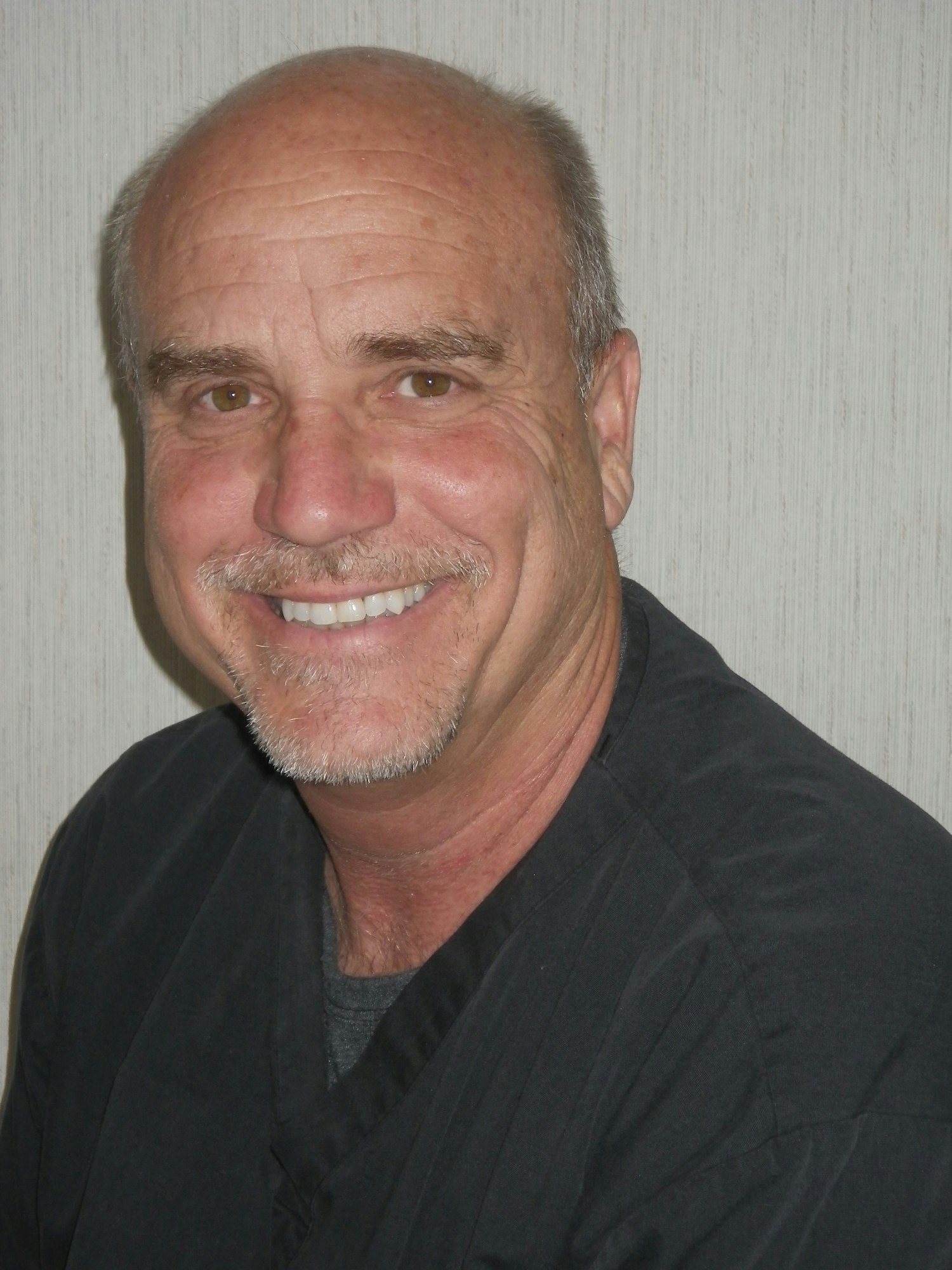 A smiling man with a mustache, wearing a black shirt, against a textured background.