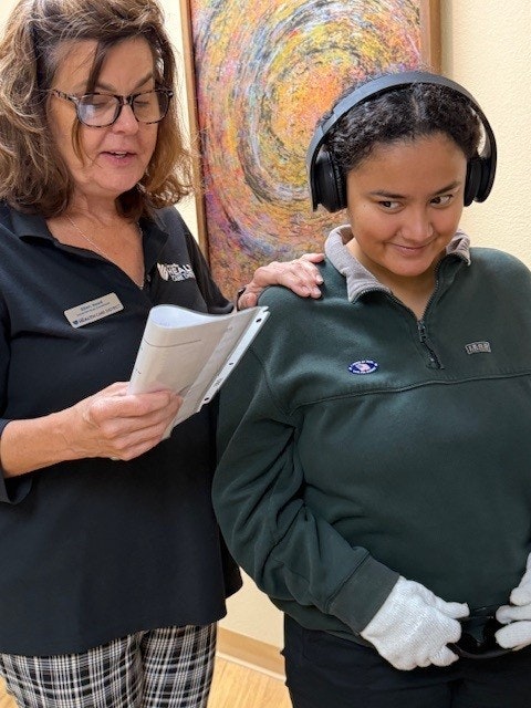 Two people: one reading a paper, the other with headphones, gloves, and a fleece, standing together with a colorful painting behind them.