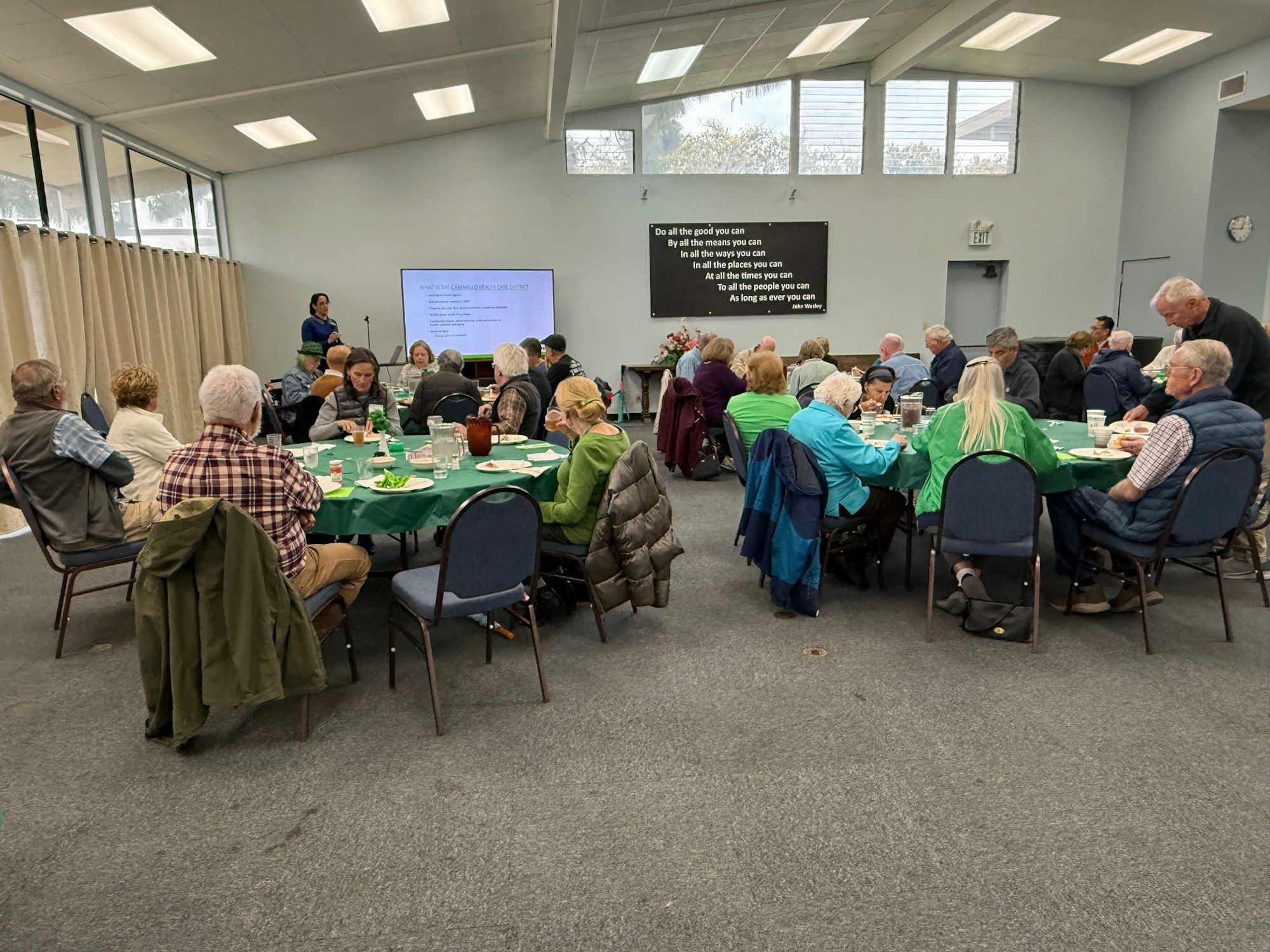 People seated at round tables in a room, listening to a speaker near a display screen.