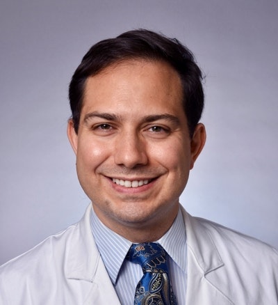 A person wearing a white coat and a tie, smiling in front of a neutral background.