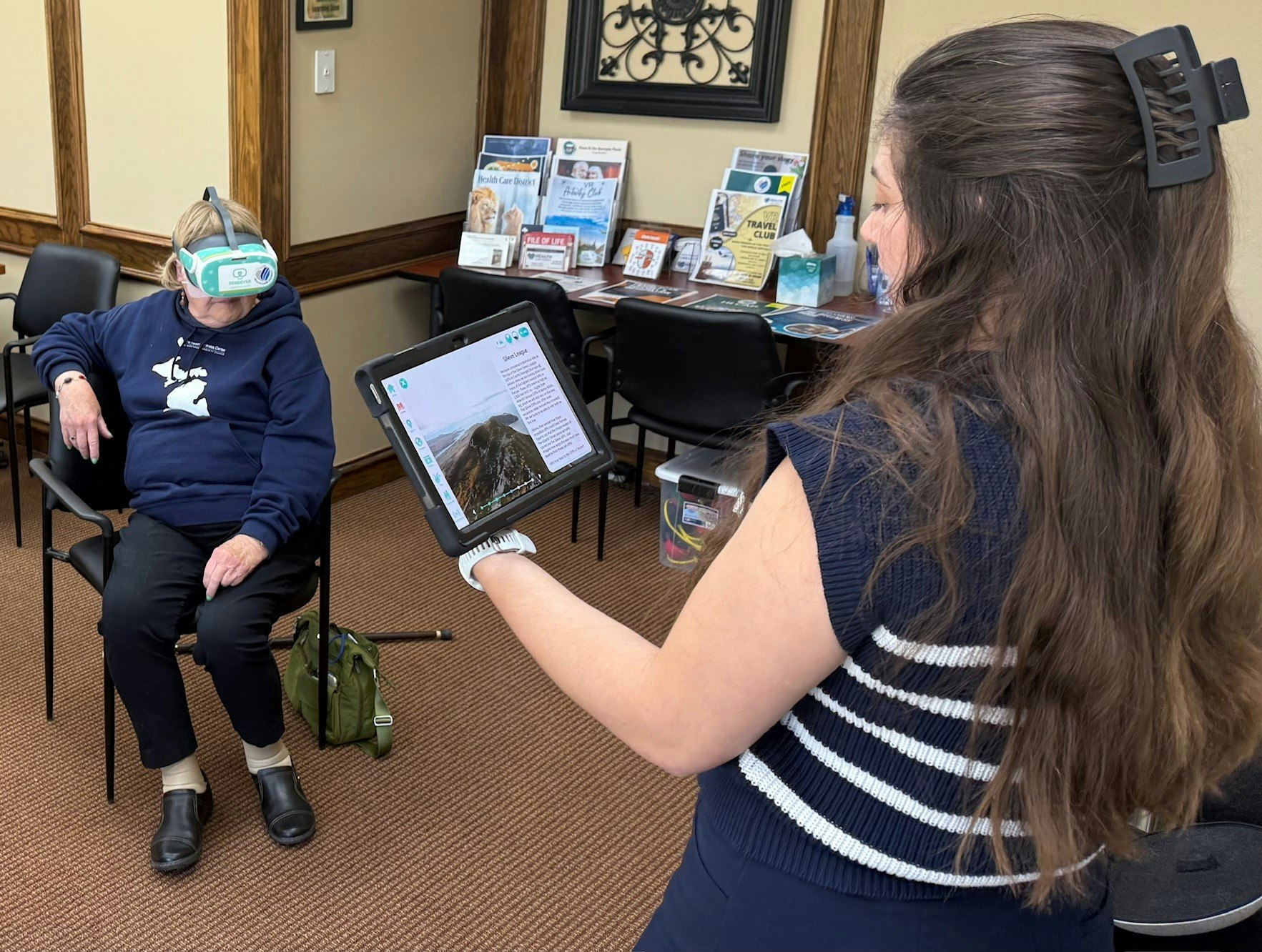 A person wearing a VR headset sits while another holds a tablet. There's a stack of brochures and decorations on a table nearby.