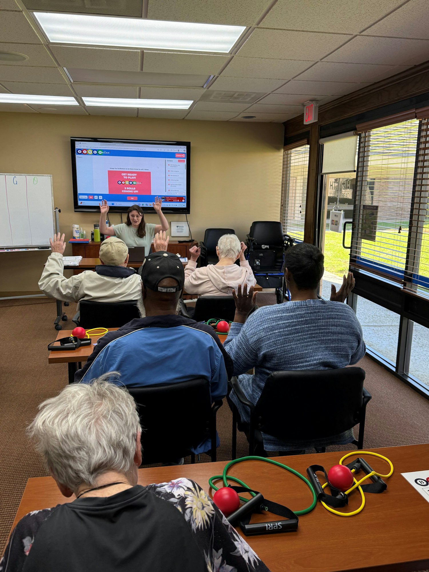A group of seniors attends a class, raising hands while a presenter explains on a screen. Exercise equipment is on the tables.