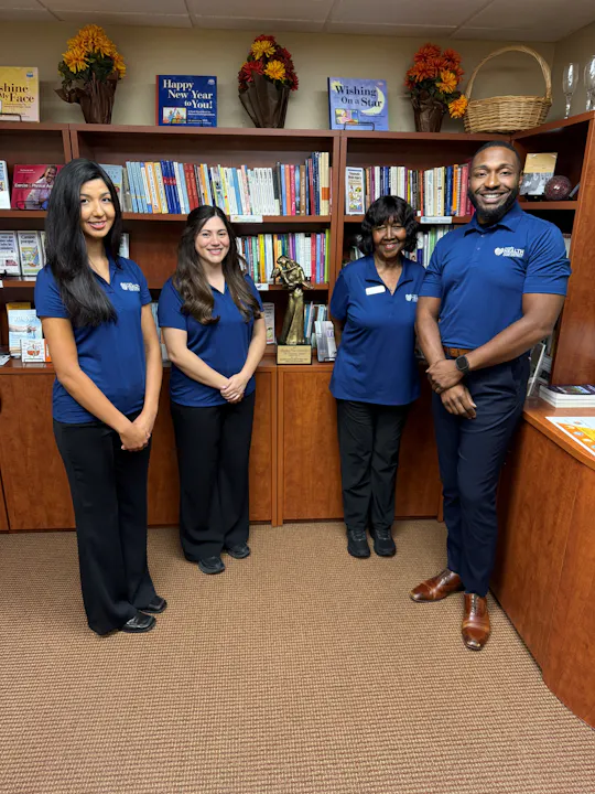 Four individuals in matching blue shirts pose for a photo in front of a bookshelf filled with books and decorations.