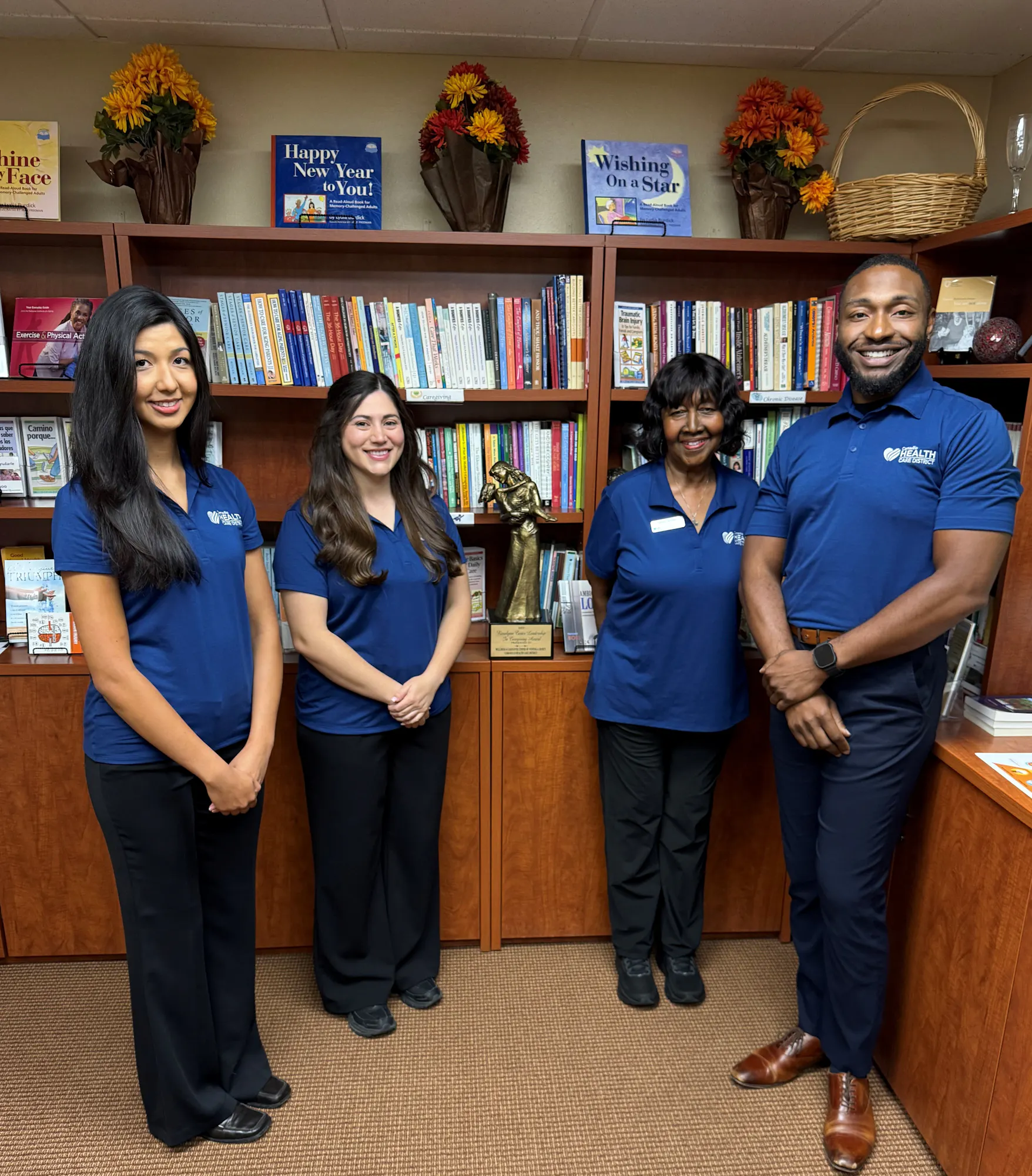 Four individuals in matching blue shirts pose for a photo in front of a bookshelf filled with books and decorations.