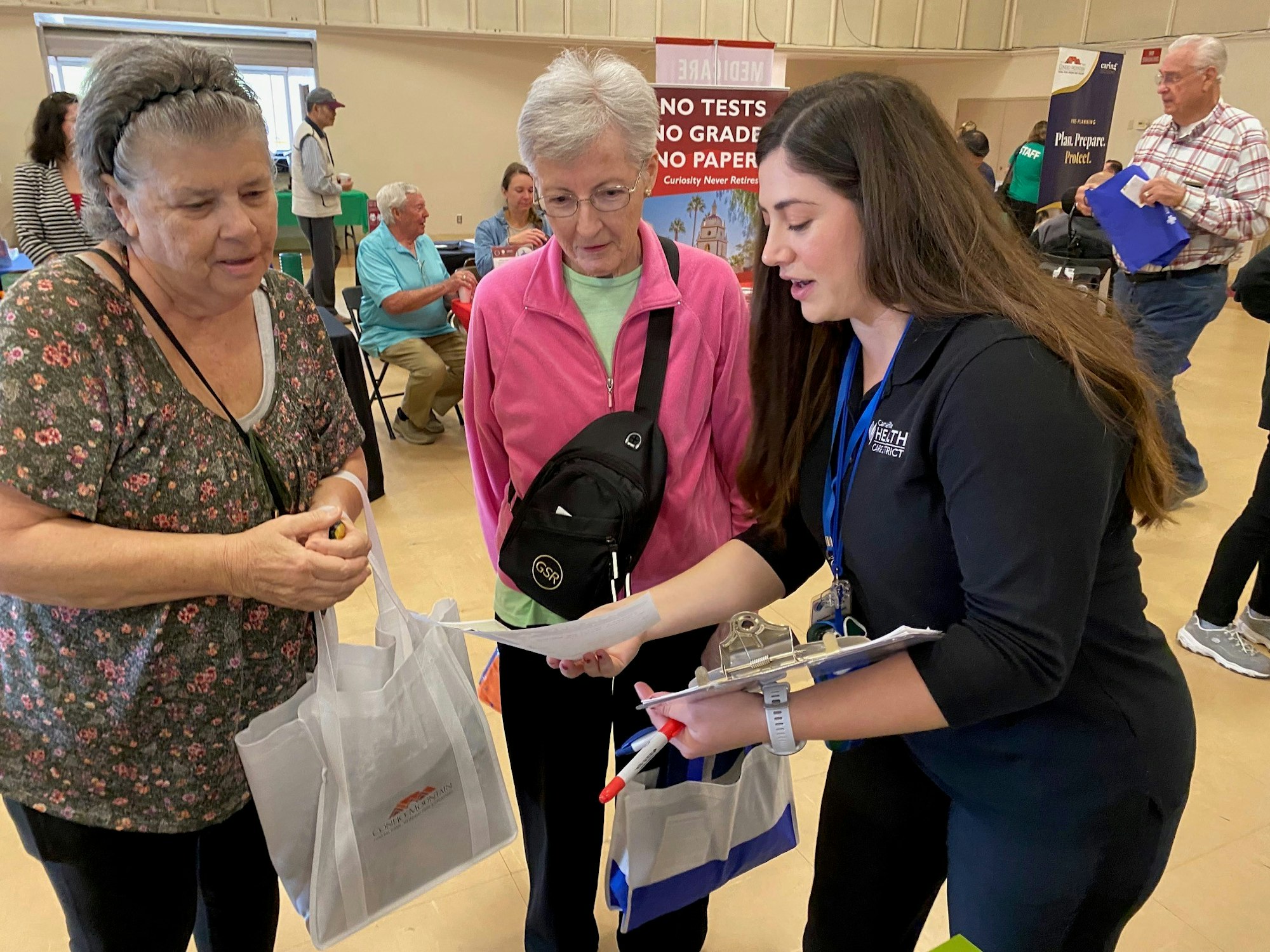 Three people interact at an event, likely informational, with one person holding a clipboard, and others holding bags.