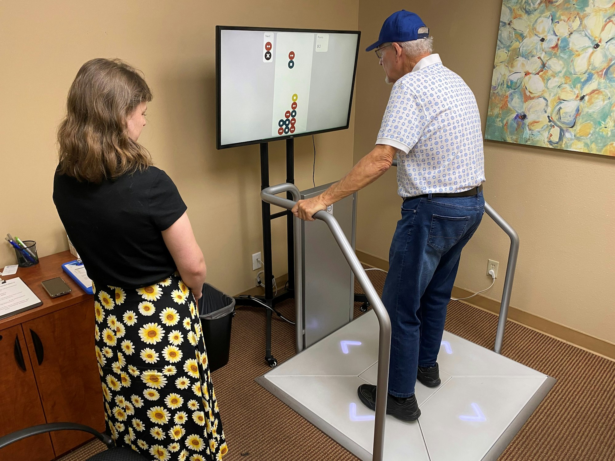 A man on a balance board interacts with a screen while a woman observes in an office setting.