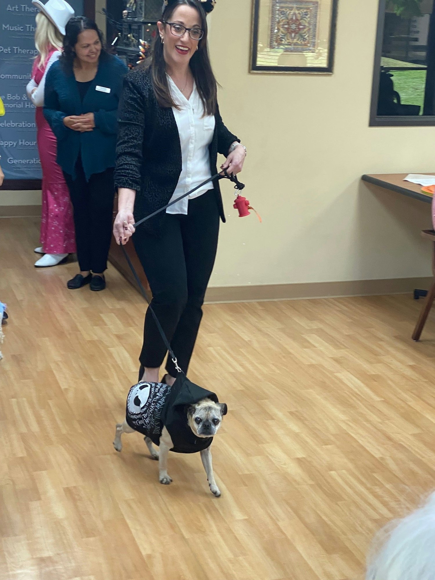A woman walks a dog wearing a hoodie indoors, with another person in the background.