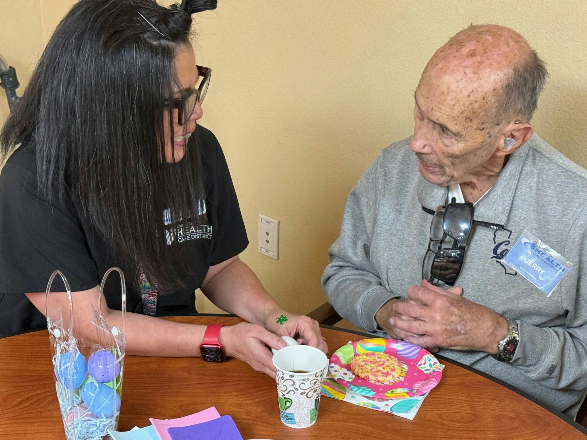 A person with long hair talks to an elderly man at a table, sharing a cup and a colorful plate with a treat.