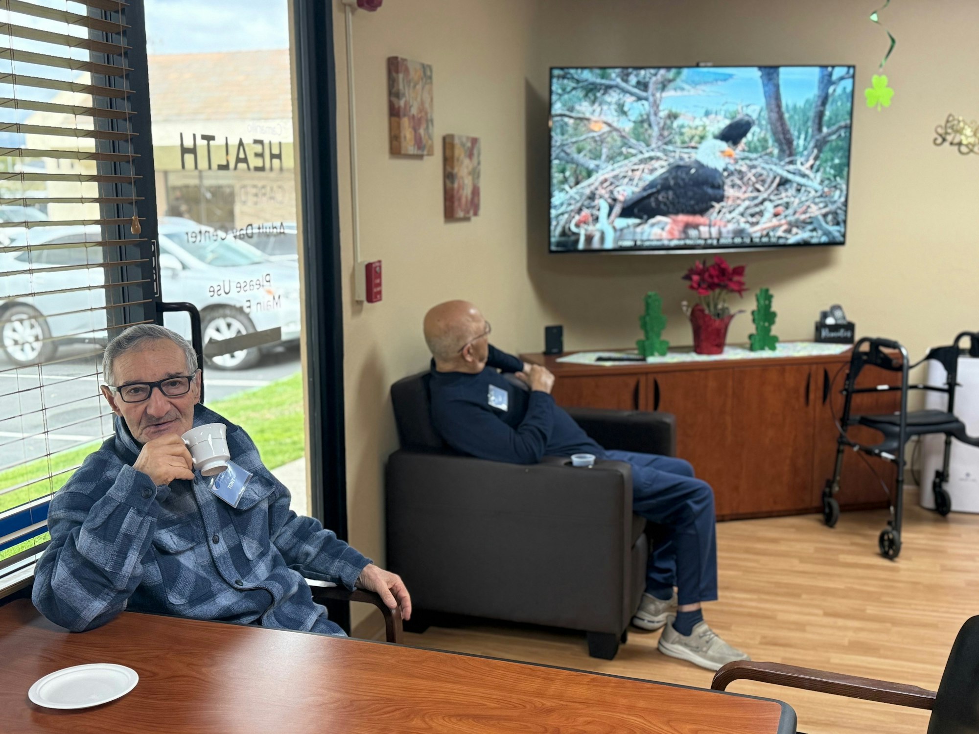 Two people in a lounge, one drinking coffee, one watching a TV with a bird on screen. A walker is nearby.