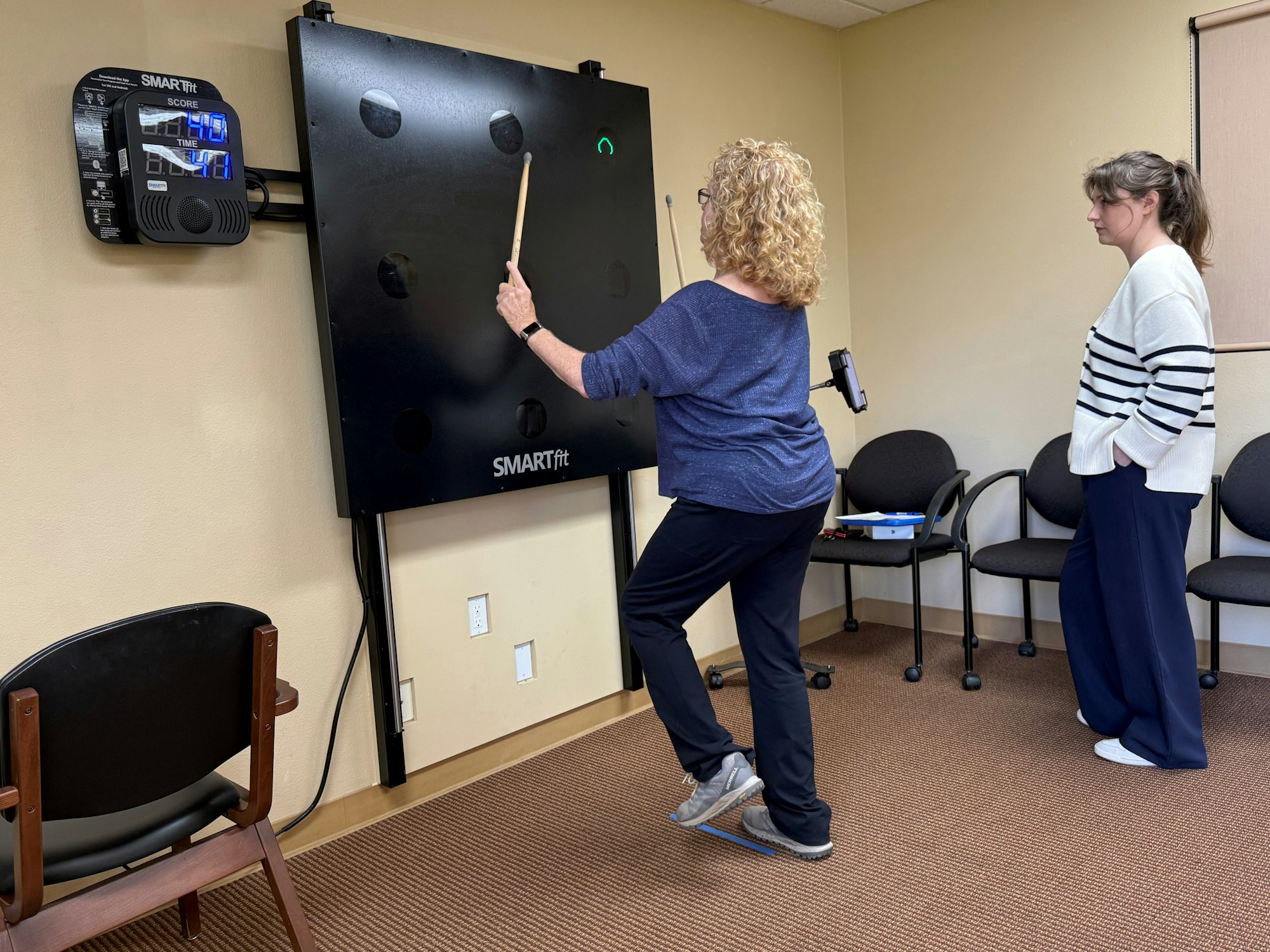 Two women are interacting with a SMARTfit interactive board in a room, one is playing while the other observes.