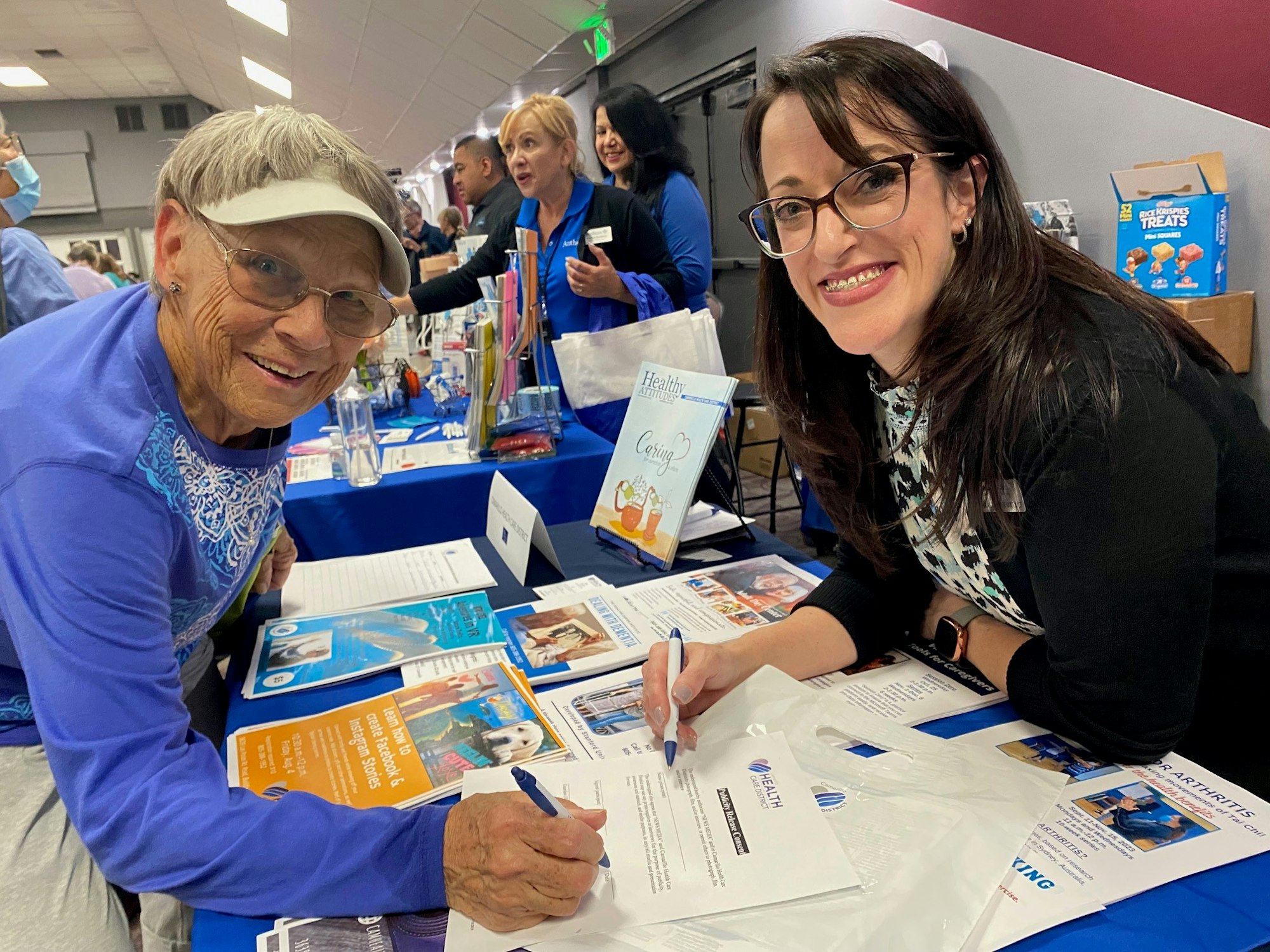 Two women at a booth with informational materials, one signing a document, in a busy event setting.
