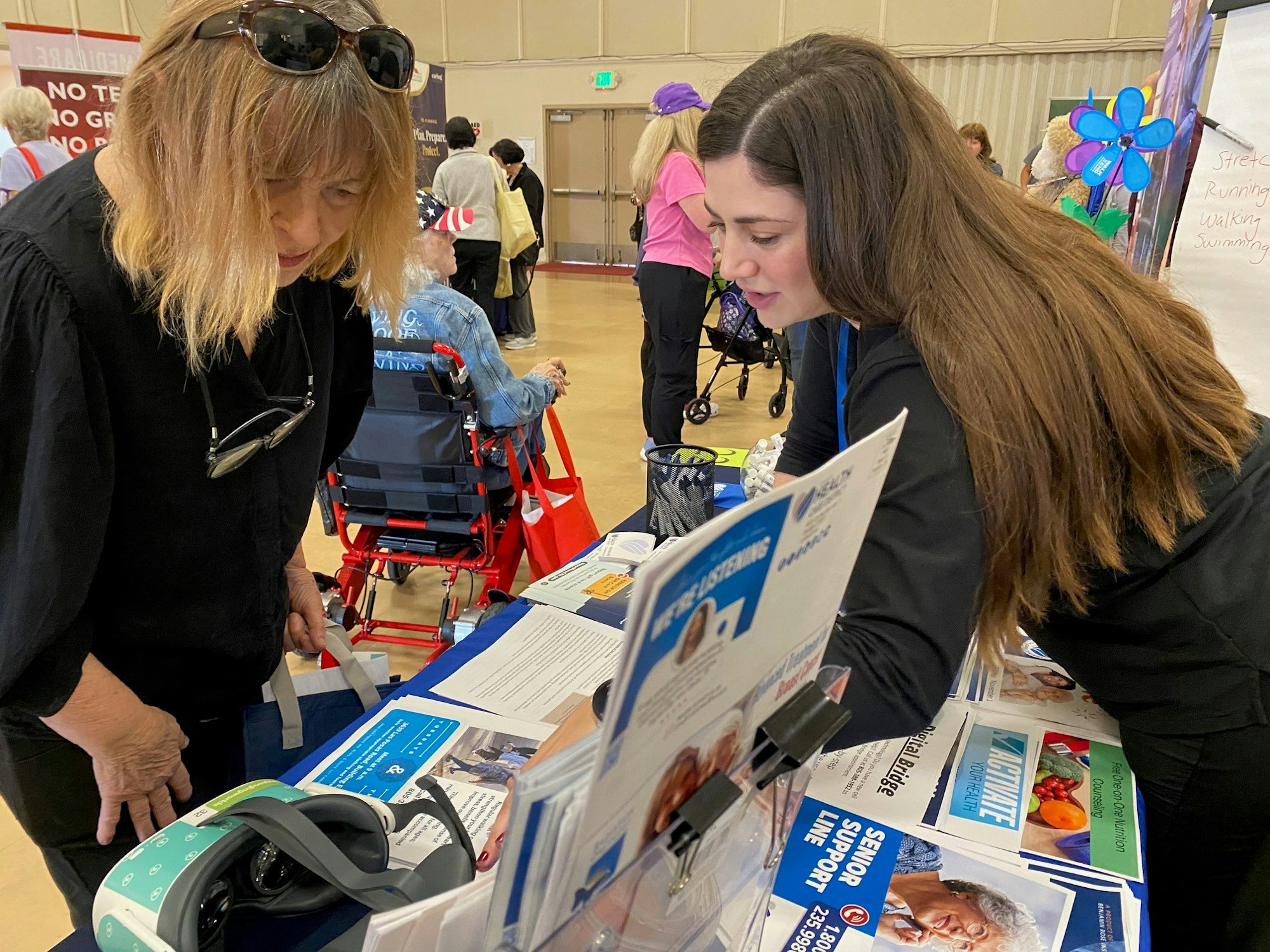 Two women at an information booth with booklets, discussing contents. Event setting visible with people in the background.