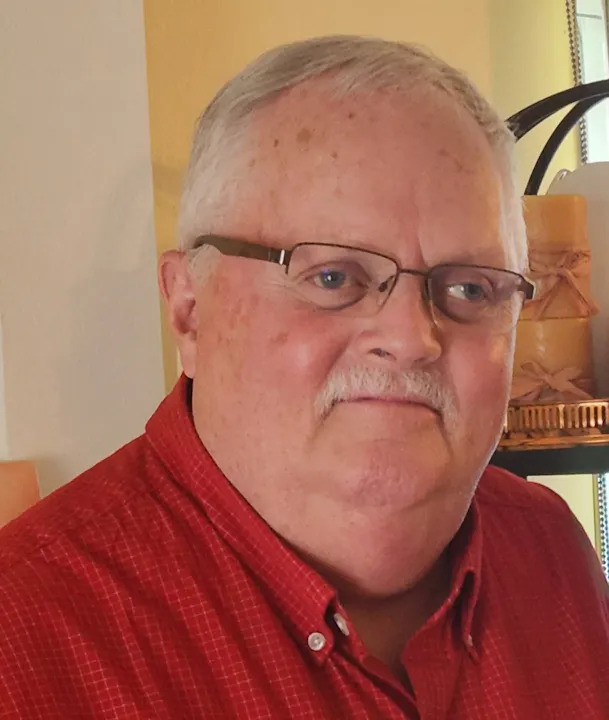 The image features a man with grey hair and glasses, wearing a red shirt, and standing indoors with decorative items in the background.