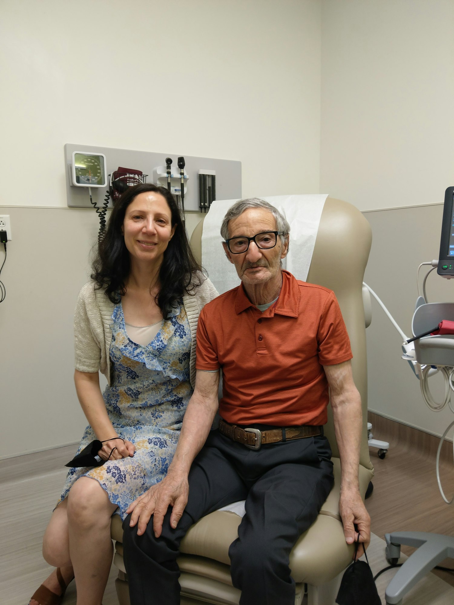 A woman and an older man sit together in a medical office, both smiling. Medical equipment is visible in the background.