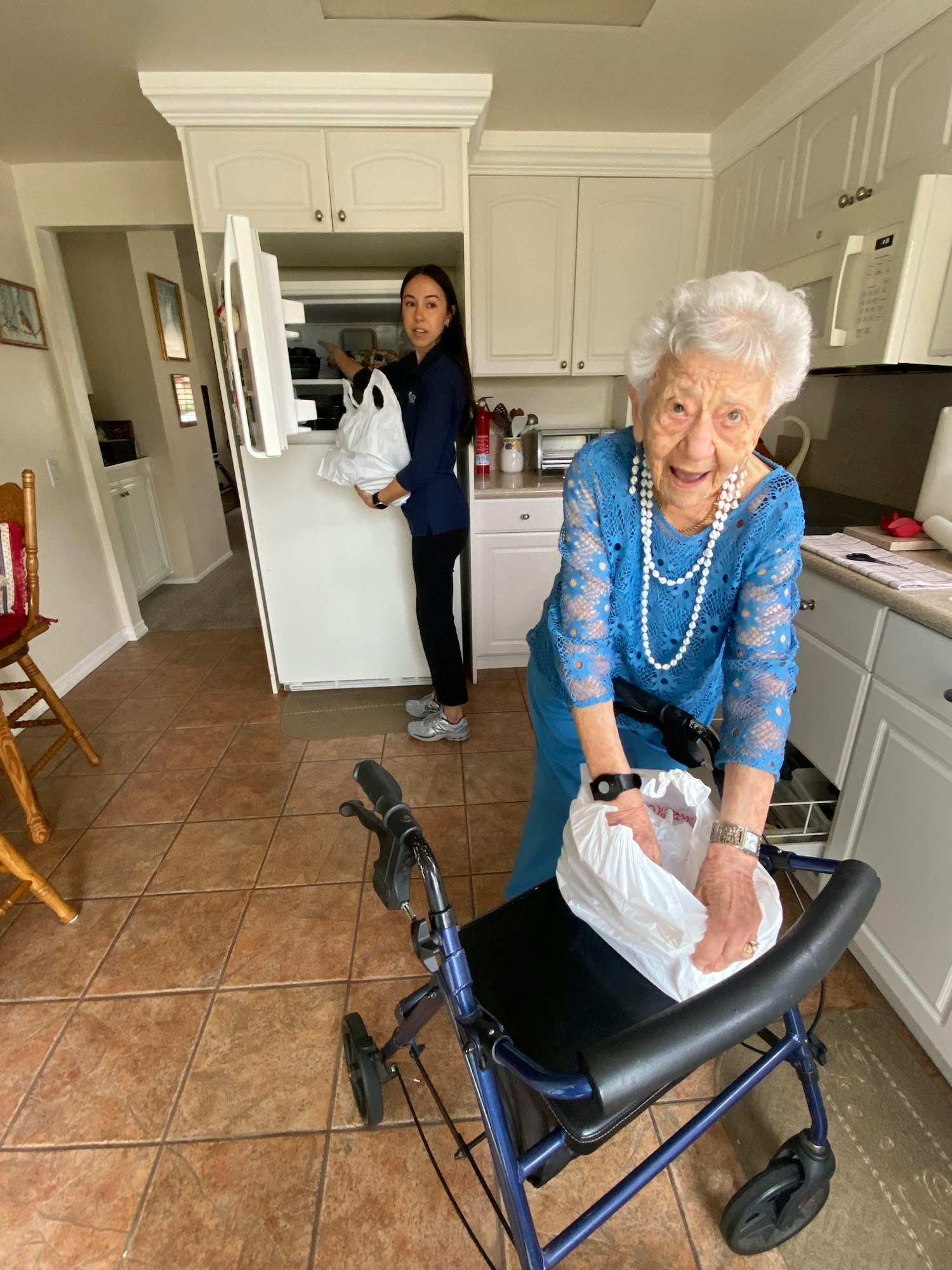 An elderly lady with a walker smiling at the camera, and a younger woman holding a bag in a kitchen.
