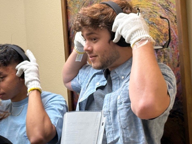 A young man adjusts headphones while wearing white gloves, standing near another person in a casual setting.