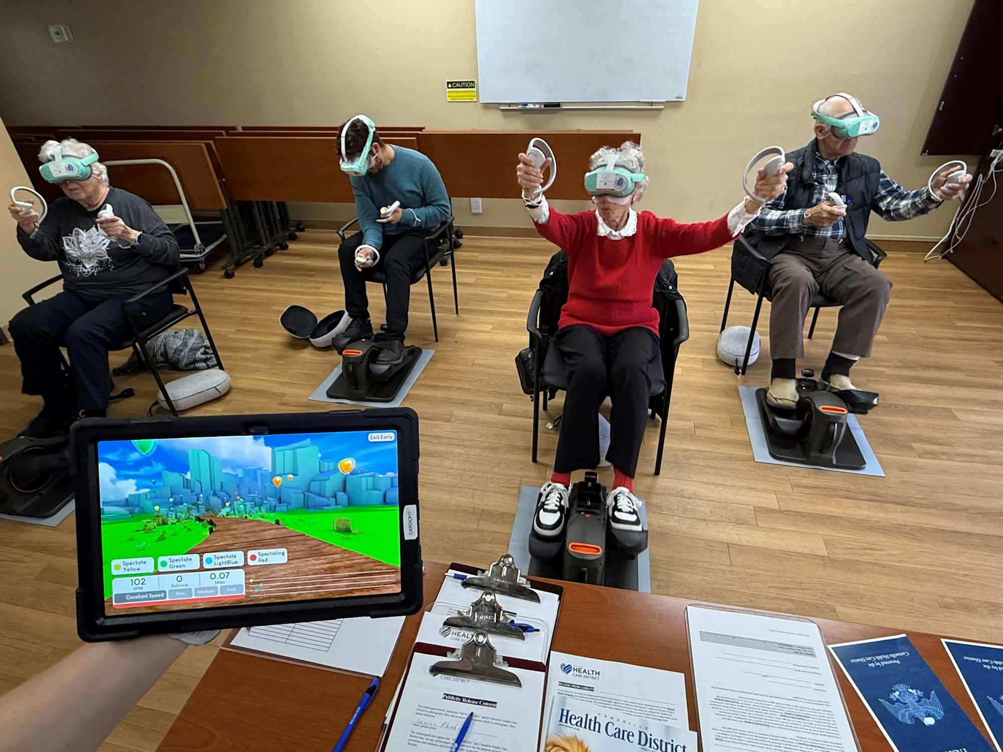 People using VR headsets seated on machines while a person holds a tablet showing a virtual environment screen.