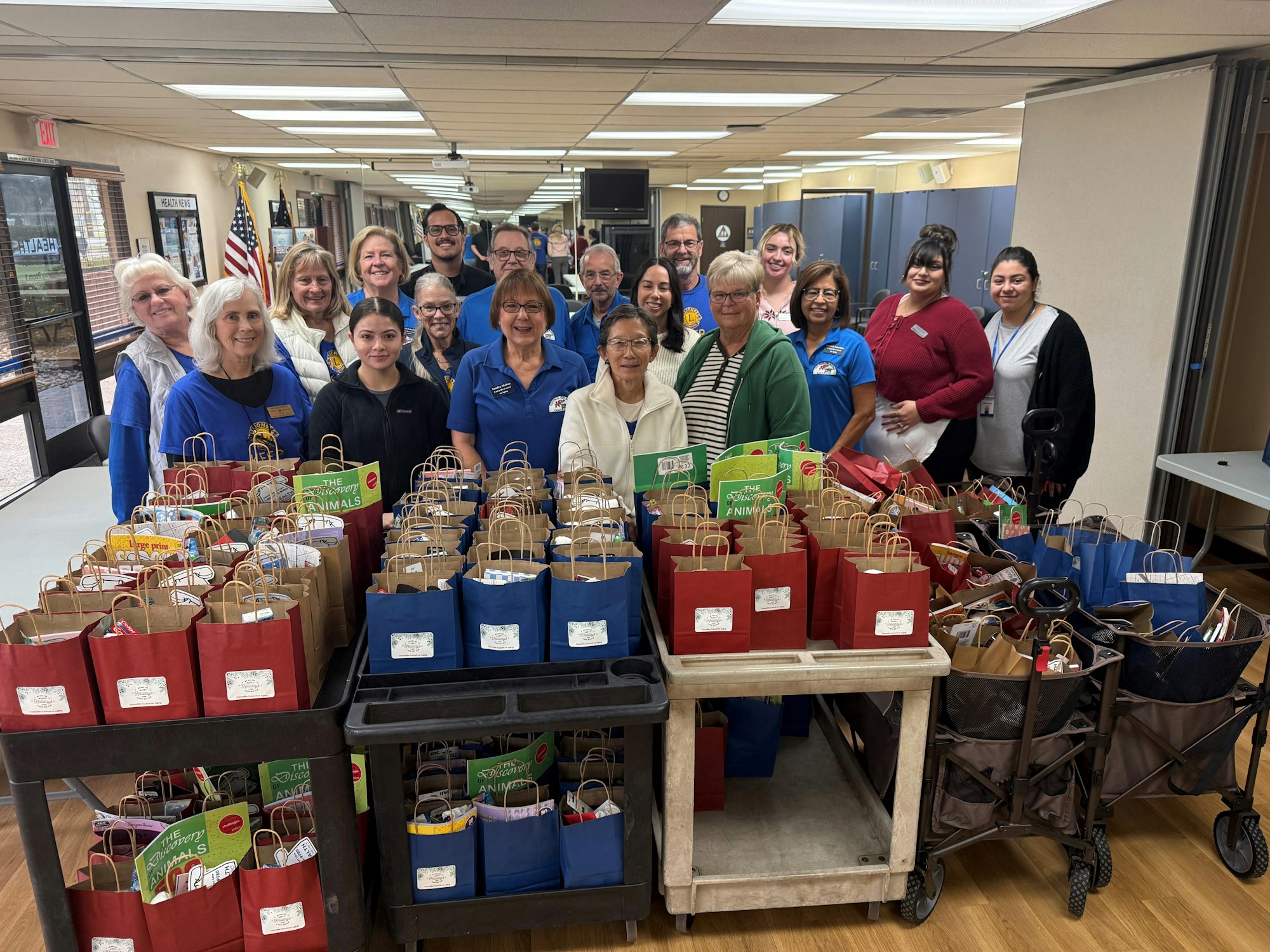 A group of people stands behind tables filled with colorful gift bags in a room with American flags.