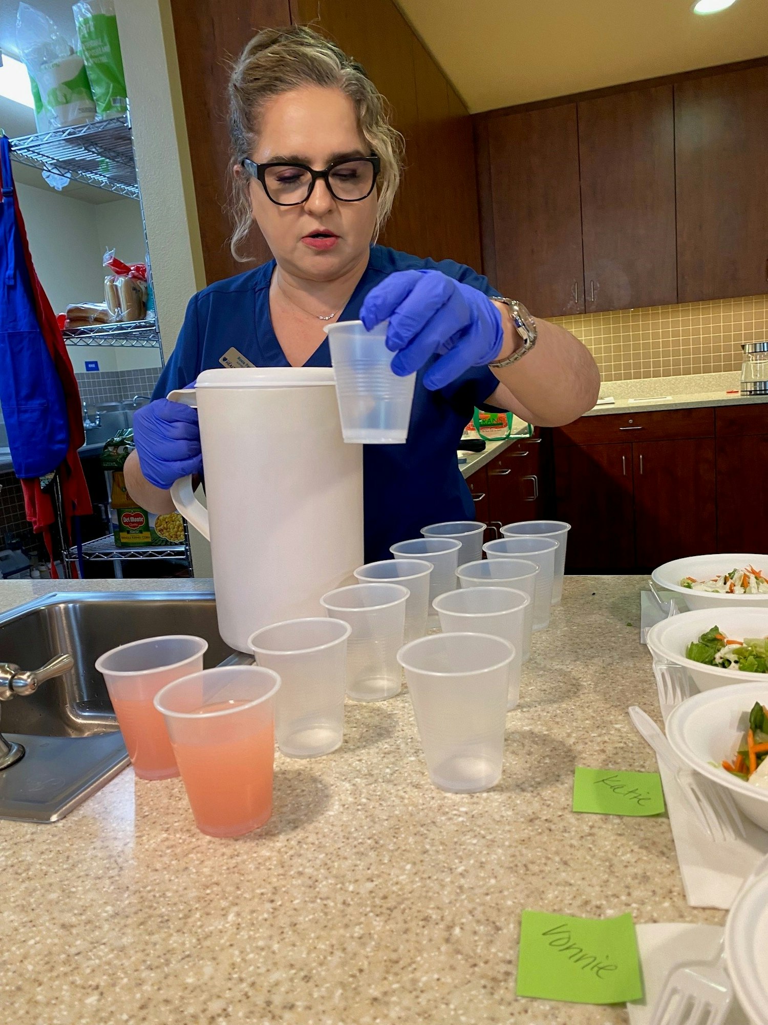 A woman in gloves pouring liquid into cups on a counter with labeled salads.