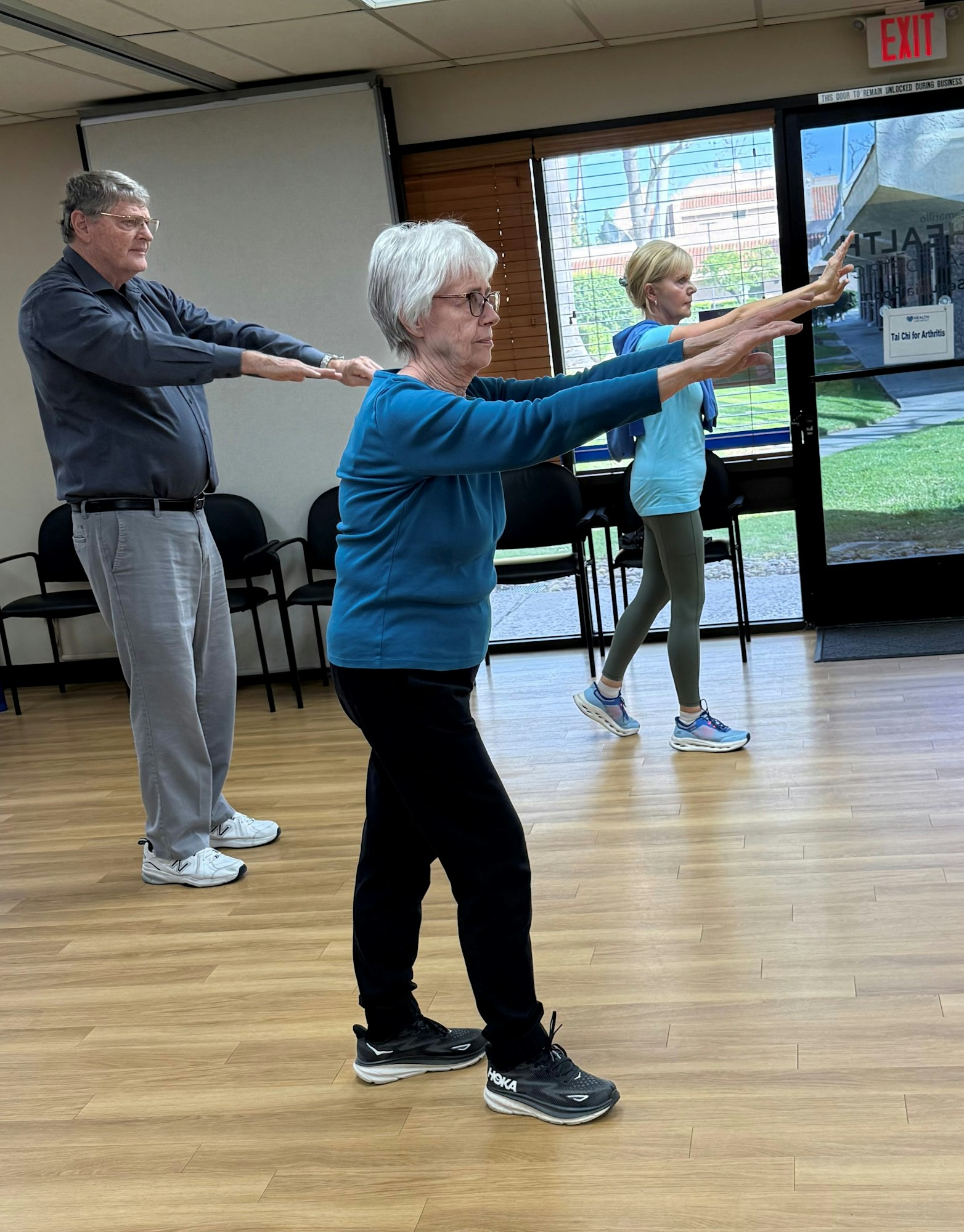Three people participate in an exercise or stretching class, extending their arms forward in a well-lit indoor setting.