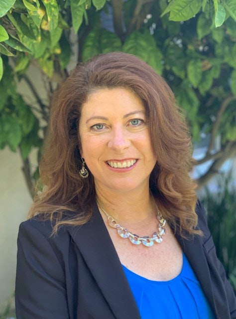 A smiling woman with brown hair, wearing a blue top and black blazer, with green foliage in the background.